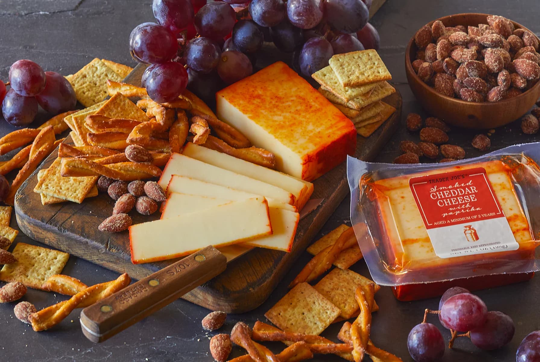 An arrangement of sliced cheddar cheese, crackers, pretzel sticks, almonds, and grapes on a wooden board, with a packaged cheddar cheese block nearby on a dark surface.
