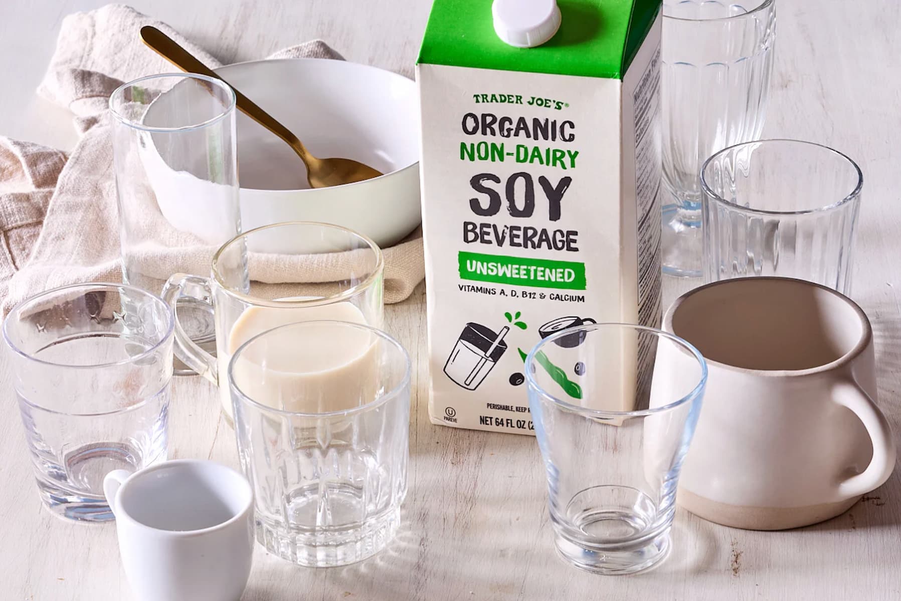 A carton of Trader Joe’s Organic Non-Dairy Soy Beverage sits on a table surrounded by several empty glasses, a mug, a bowl with a spoon, and a folded napkin. One glass is filled with the soy beverage.