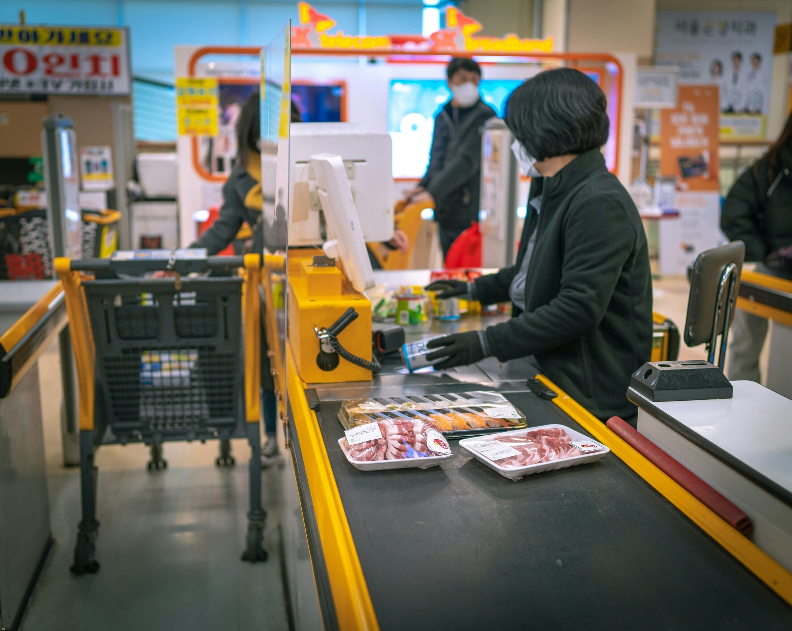 A supermarket cashier scans groceries, including packaged meat and sushi, while a customer waits. Other shoppers are visible in the background. The scene takes place at a modern checkout counter.