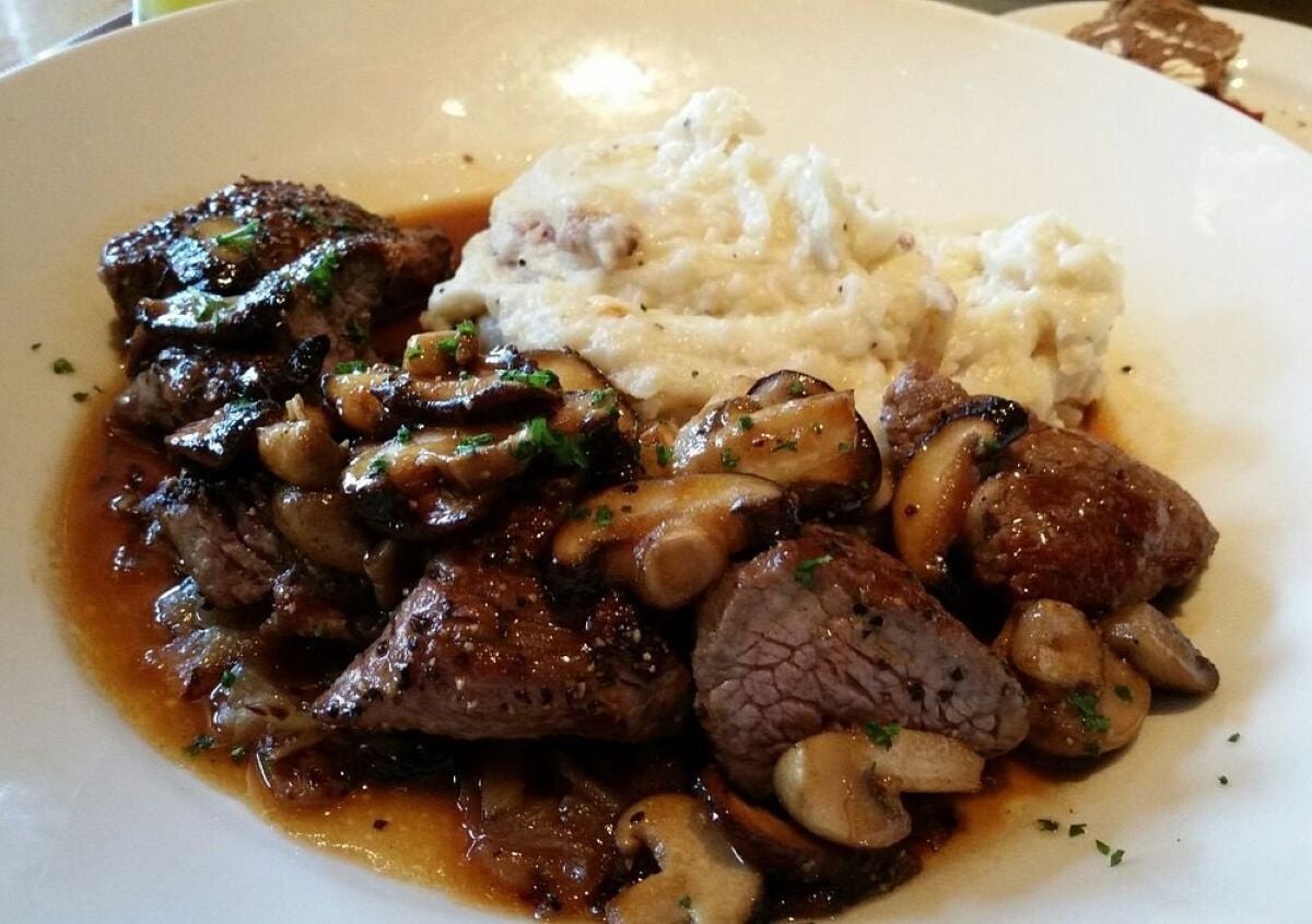 A plate of beef tips cooked with mushrooms in a brown sauce, served alongside a generous portion of mashed potatoes.