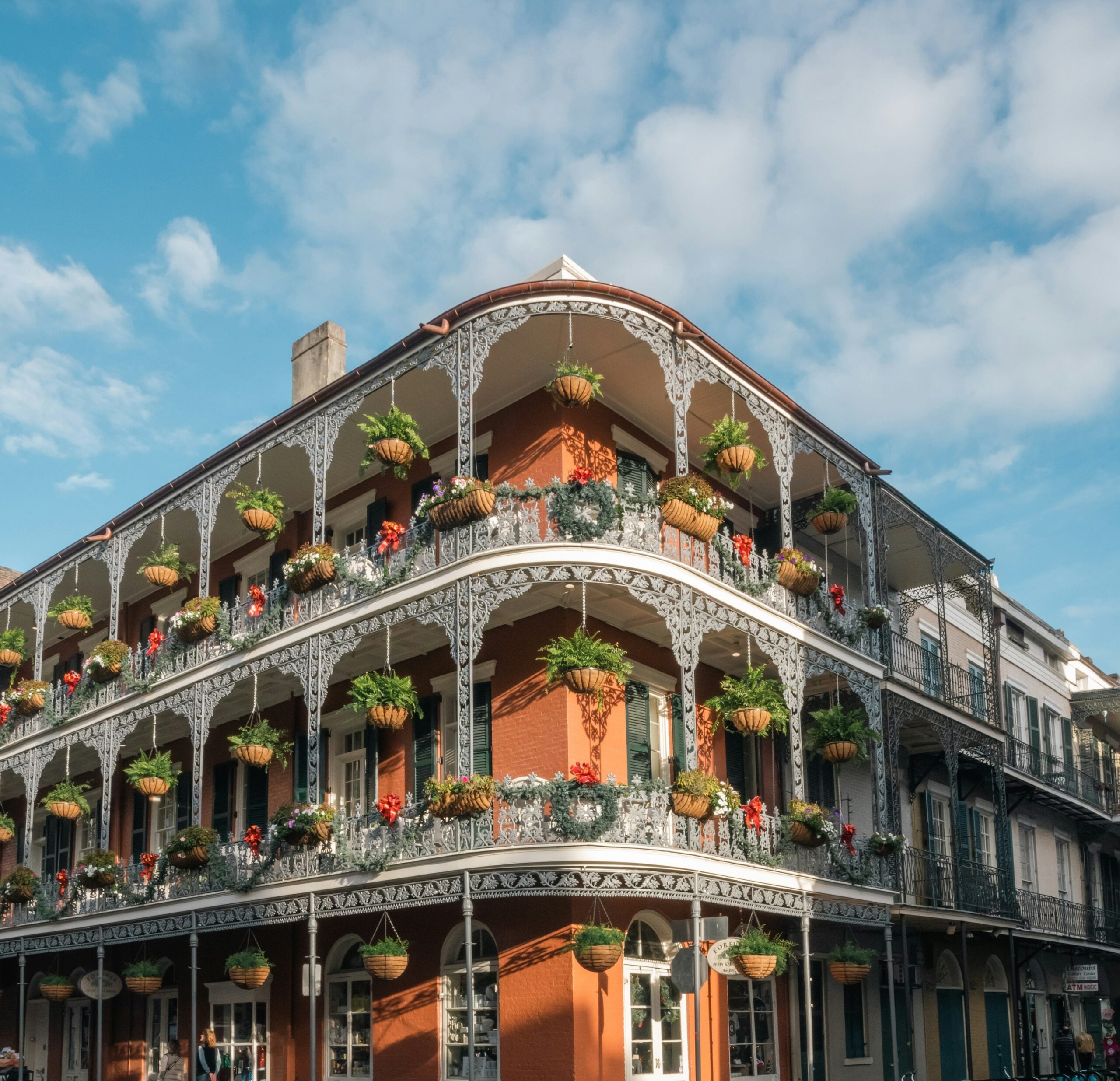 A corner building in New Orleans’ French Quarter with ornate iron balconies decorated with hanging flower baskets, red brick walls, and blue sky with scattered clouds in the background.