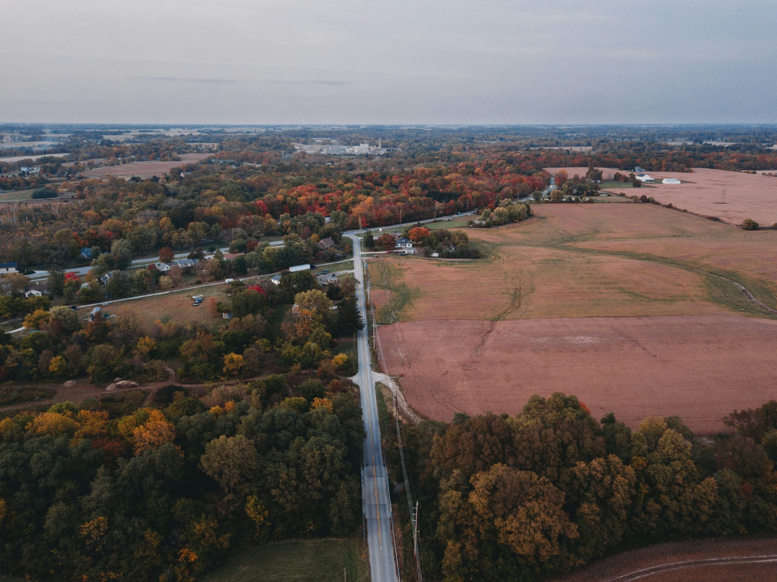 Aerial view of a rural landscape with a road dividing farmland, scattered trees, and houses. Fields appear brown and green, with trees showing autumn colors under a cloudy sky.