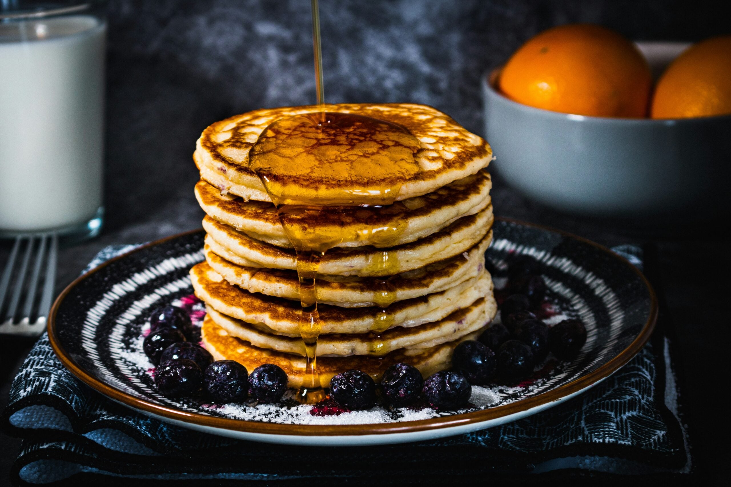 A stack of pancakes topped with syrup, served on a dark plate with fresh blueberries and powdered sugar, with a bowl of oranges and a glass of milk in the background.