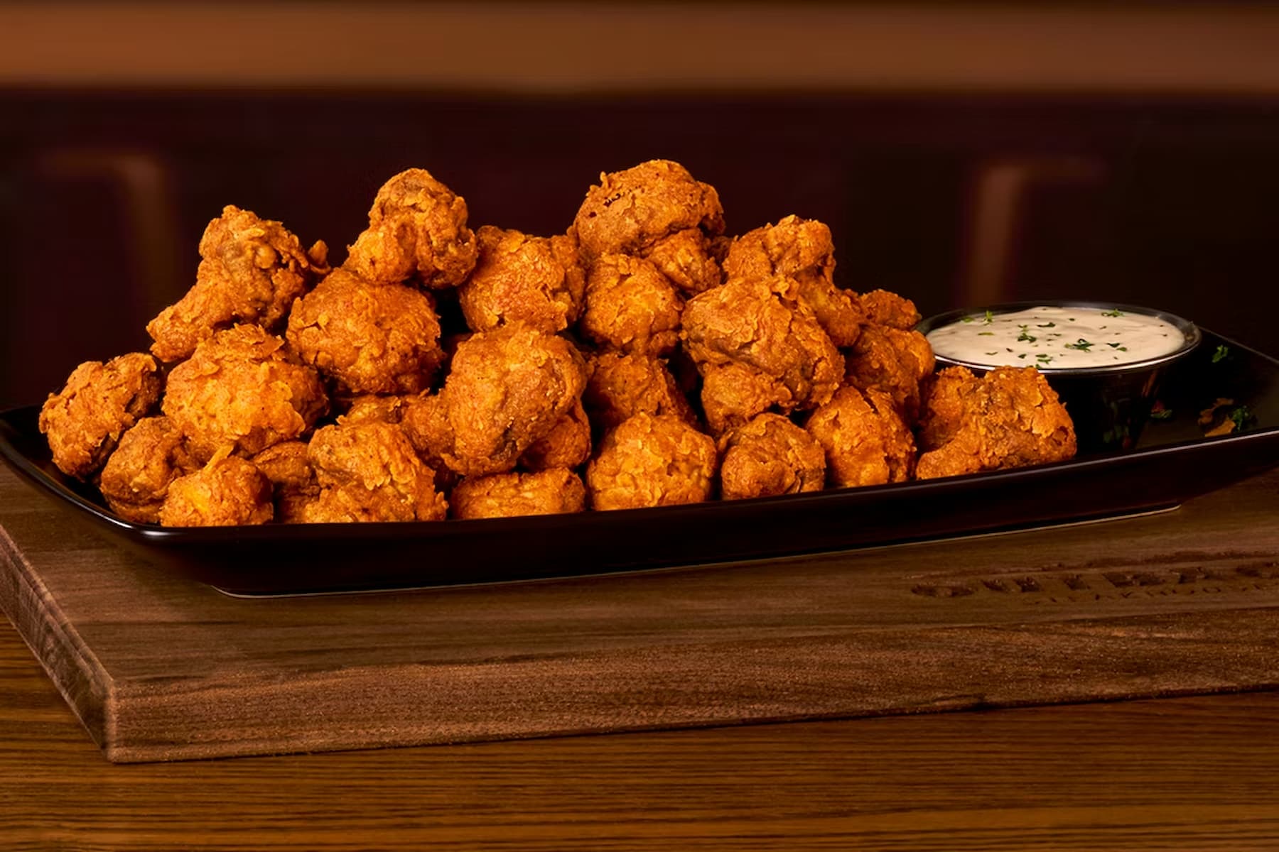 A black rectangular plate filled with crispy fried chicken wings, served with a side of creamy dipping sauce garnished with herbs, placed on a wooden surface.