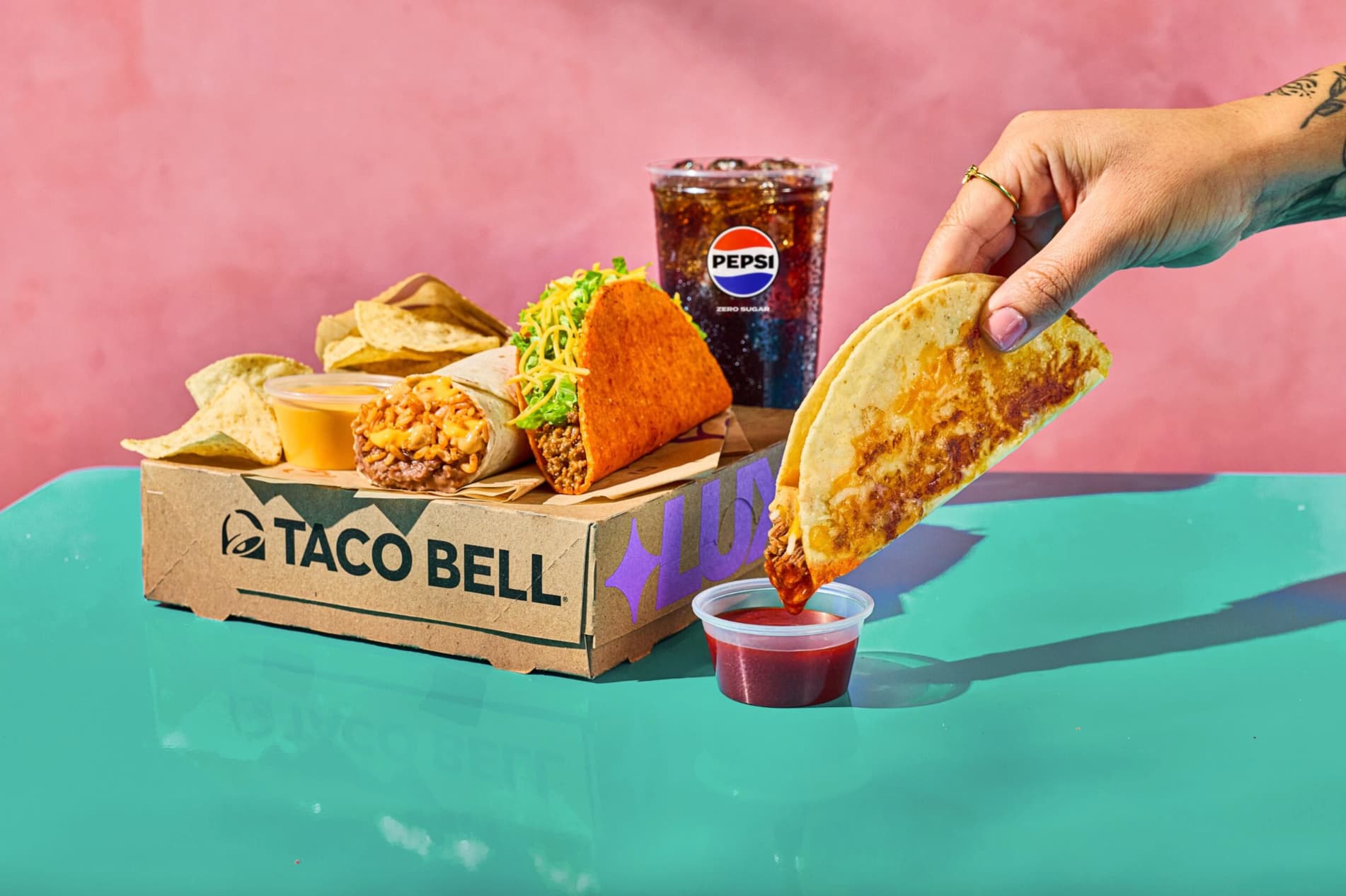 A hand dips a Taco Bell quesadilla into sauce beside a box with nachos, taco, cheesy crunch wrap, and a cup of Pepsi on a turquoise table against a pink background.