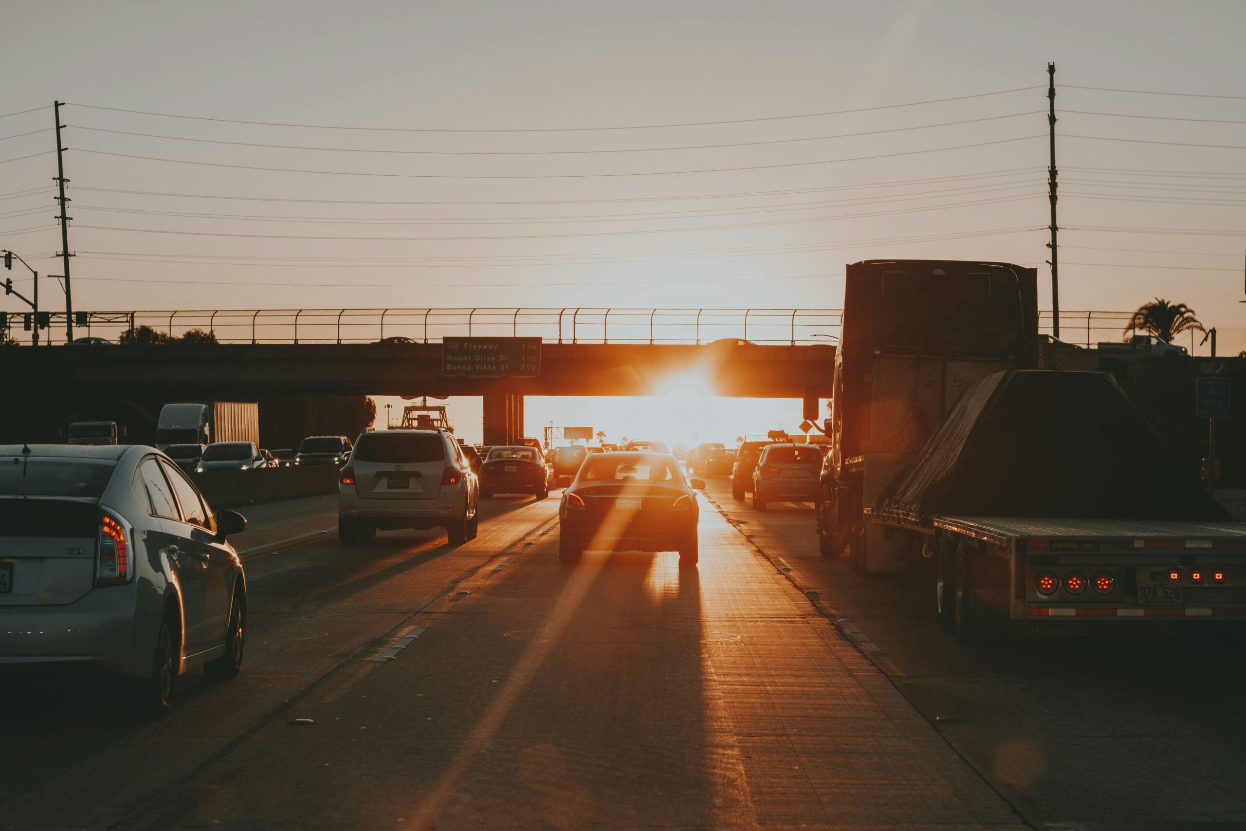 Cars driving on a busy highway at sunset, with the sun shining brightly above the horizon and casting long shadows, an overpass in the background, and vehicles traveling in both directions.