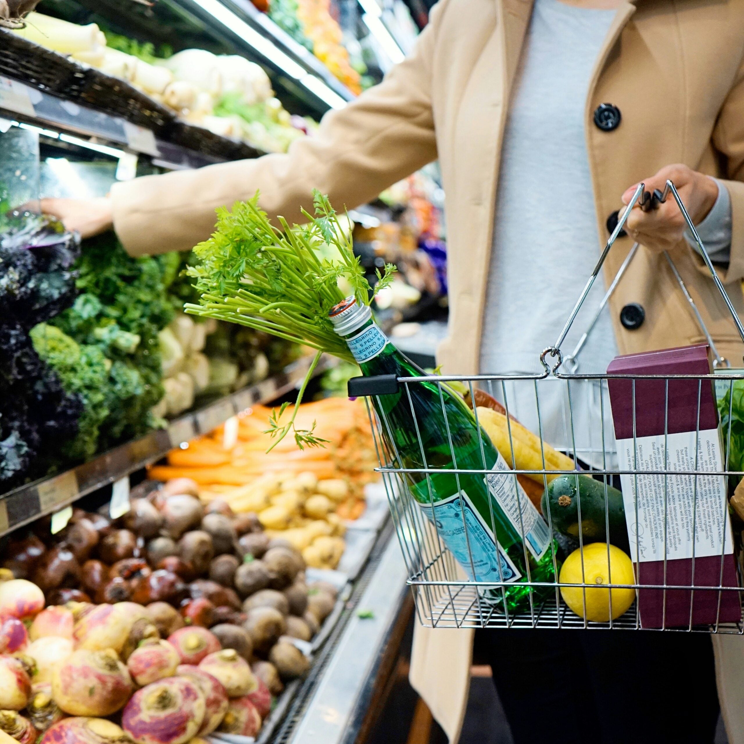 A person wearing a beige coat shops for fresh produce, reaching for vegetables while holding a basket filled with greens, bottled water, a lemon, and packaged goods in a grocery store aisle.