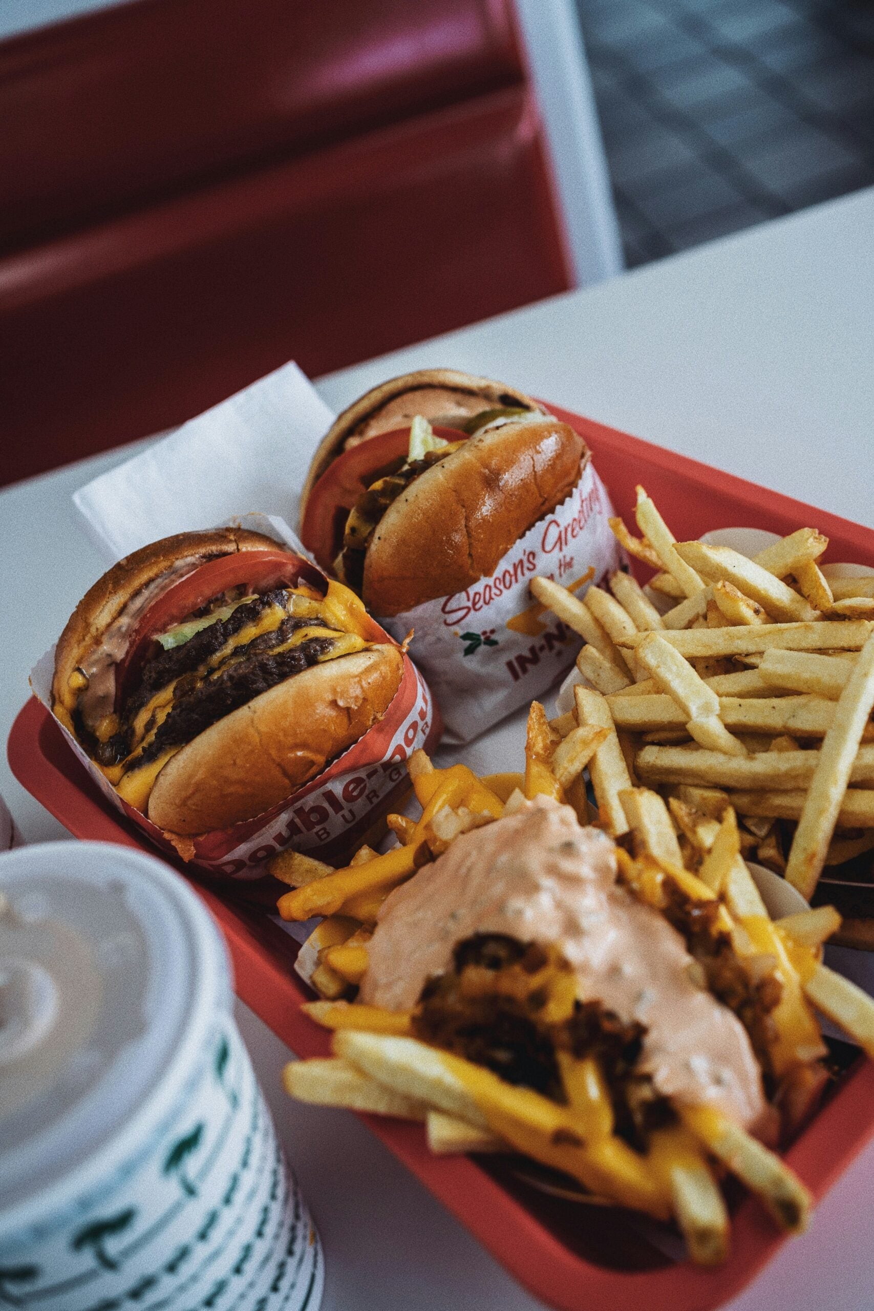 A red tray holds two cheeseburgers with lettuce, tomato, and onion, a serving of regular fries, a serving of loaded fries with sauce and grilled onions, and a soft drink in a white cup.