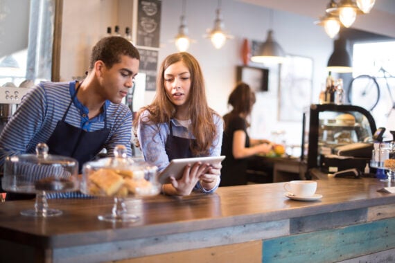 Two teens working summer jobs as baristas stand behind a caf&eacute; counter, wearing aprons and looking at a tablet together. Pastries sit in glass stands, and a cup of coffee is on the counter. Another worker is visible in the background.