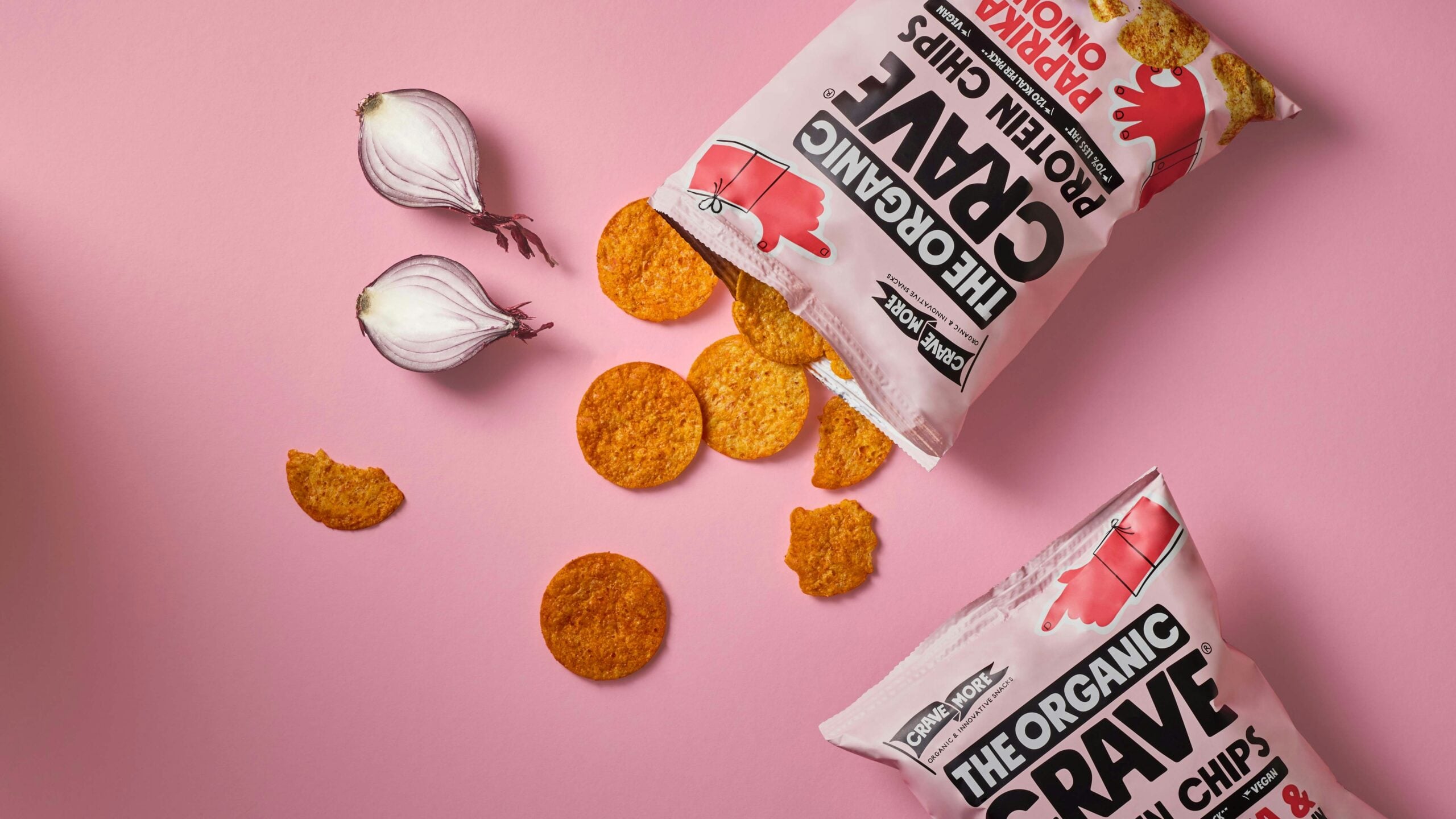 Two bags of The Organic Crave protein chips, a halved red onion, and several round chips scattered on a pink background.