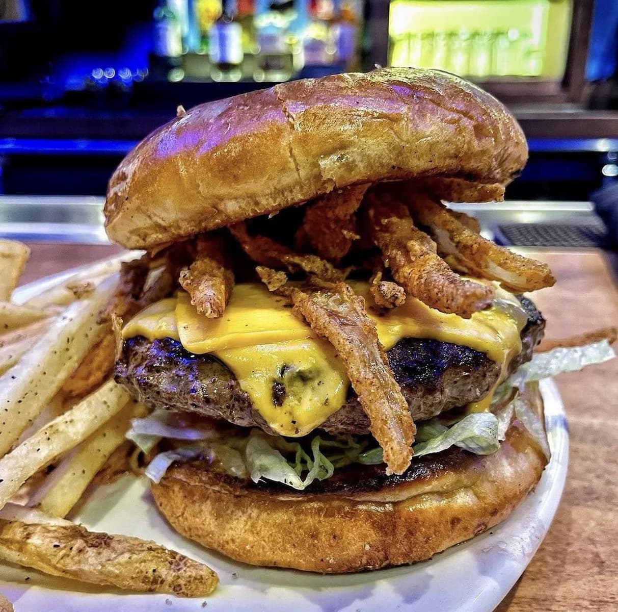 A close-up of a cheeseburger with crispy fried onions, melted cheddar cheese, lettuce, and a toasted bun, served with a side of seasoned French fries on a white plate. A bar setup is blurred in the background.