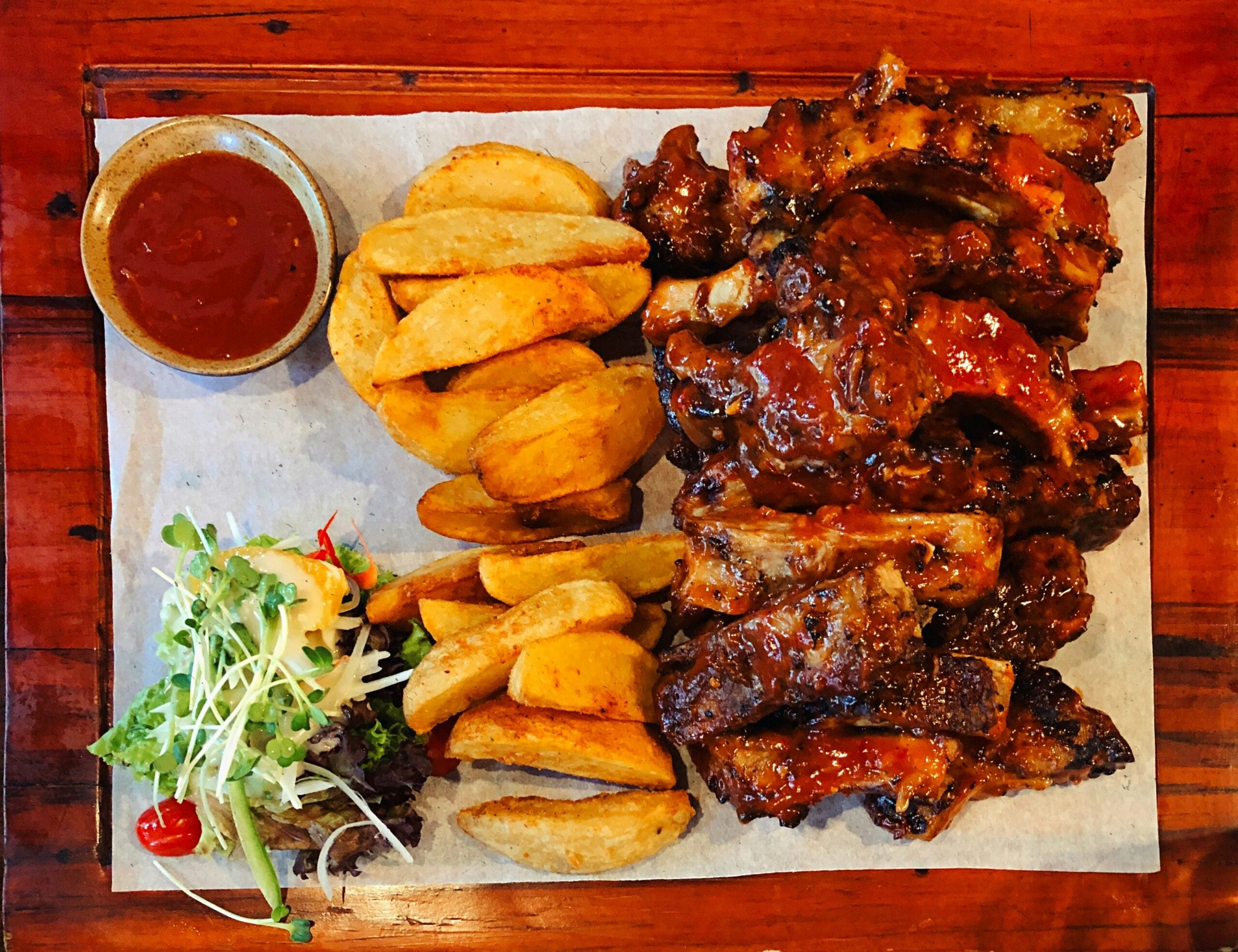 A serving of glazed BBQ ribs, crispy potato wedges, a side of dipping sauce, and a small salad with greens and cherry tomatoes on a wooden table.