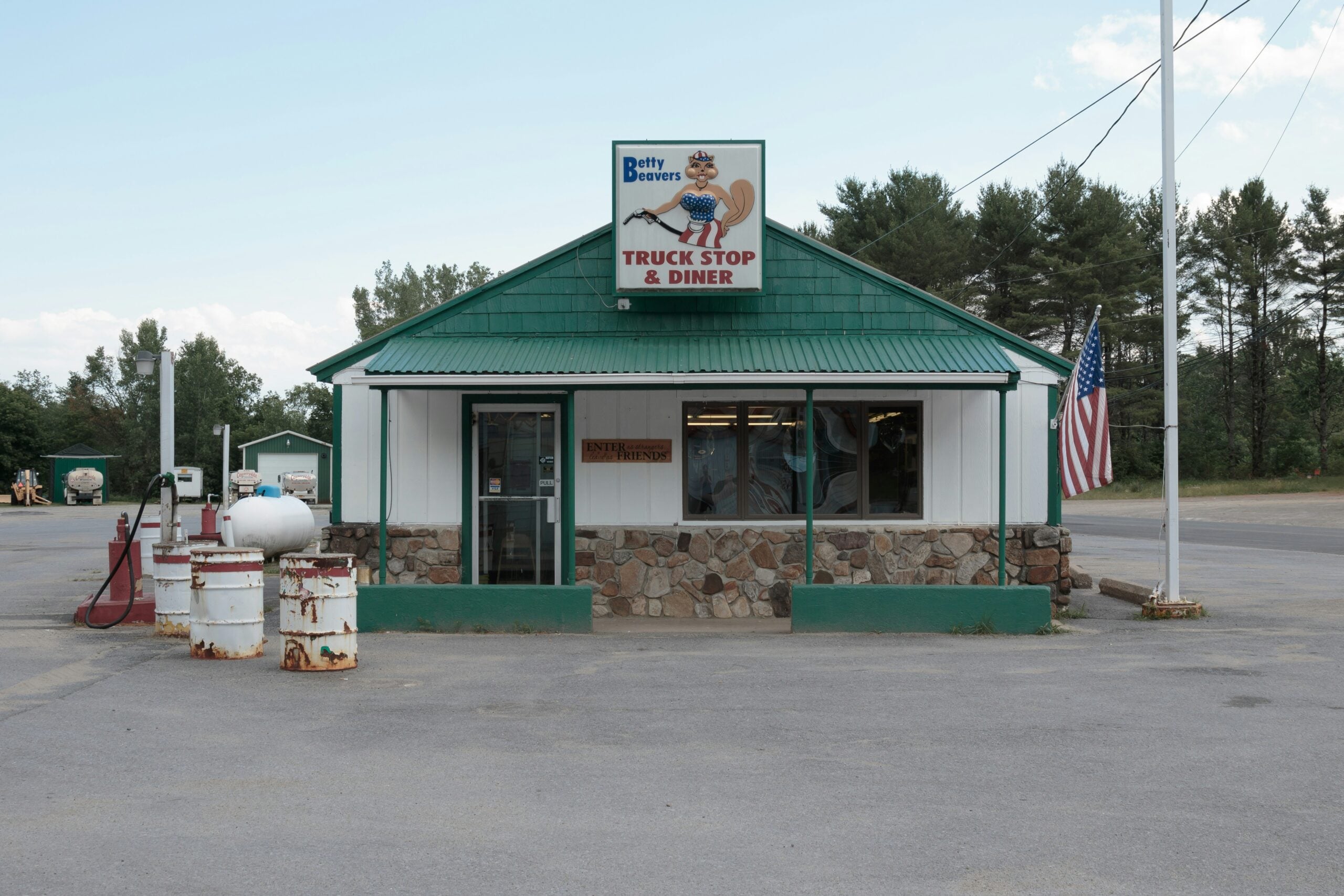 A small roadside building with a green roof and a sign reading "Betty Beavers Truck Stop & Diner." Old gas pumps are out front, and an American flag is displayed by the entrance. Pine trees are in the background.