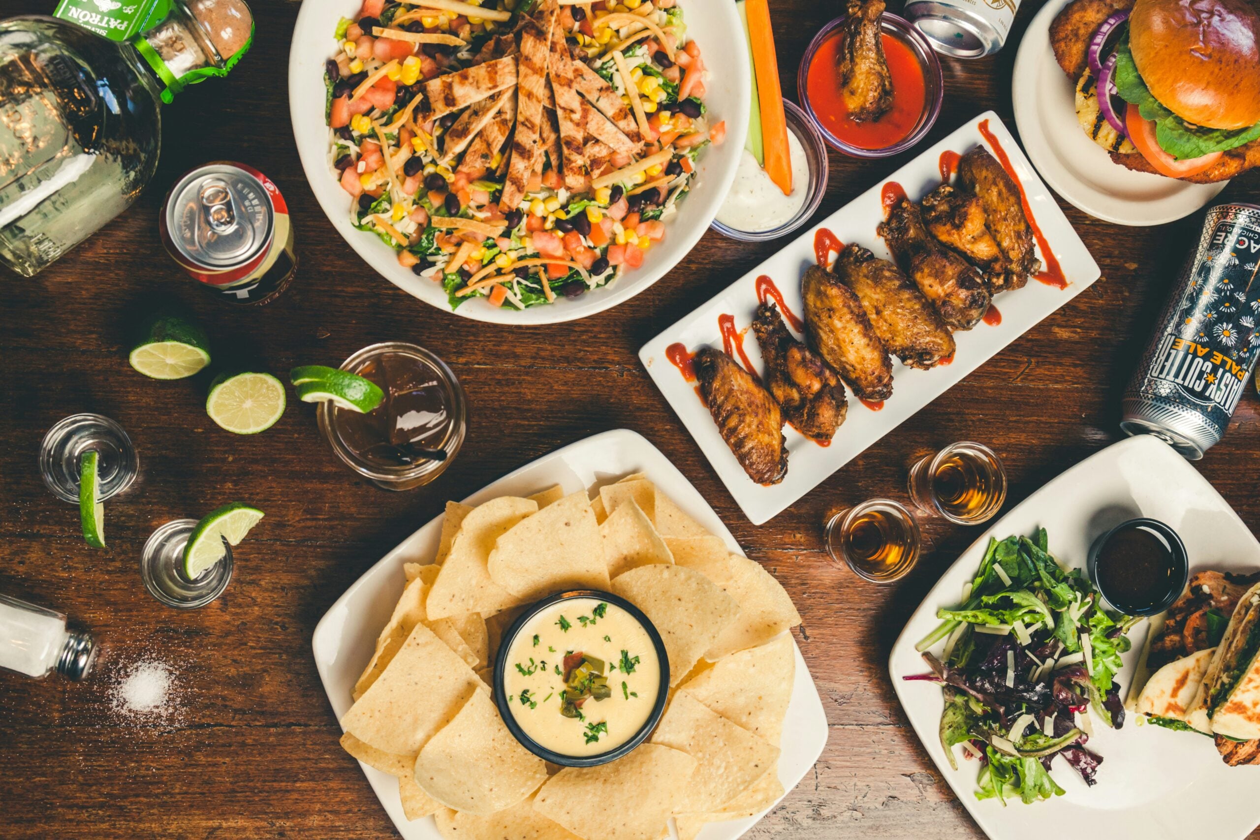 An overhead view of a table with grilled chicken salad, nachos with cheese dip, chicken wings, mixed greens, burgers, and drinks including soda, beer, and cocktails.