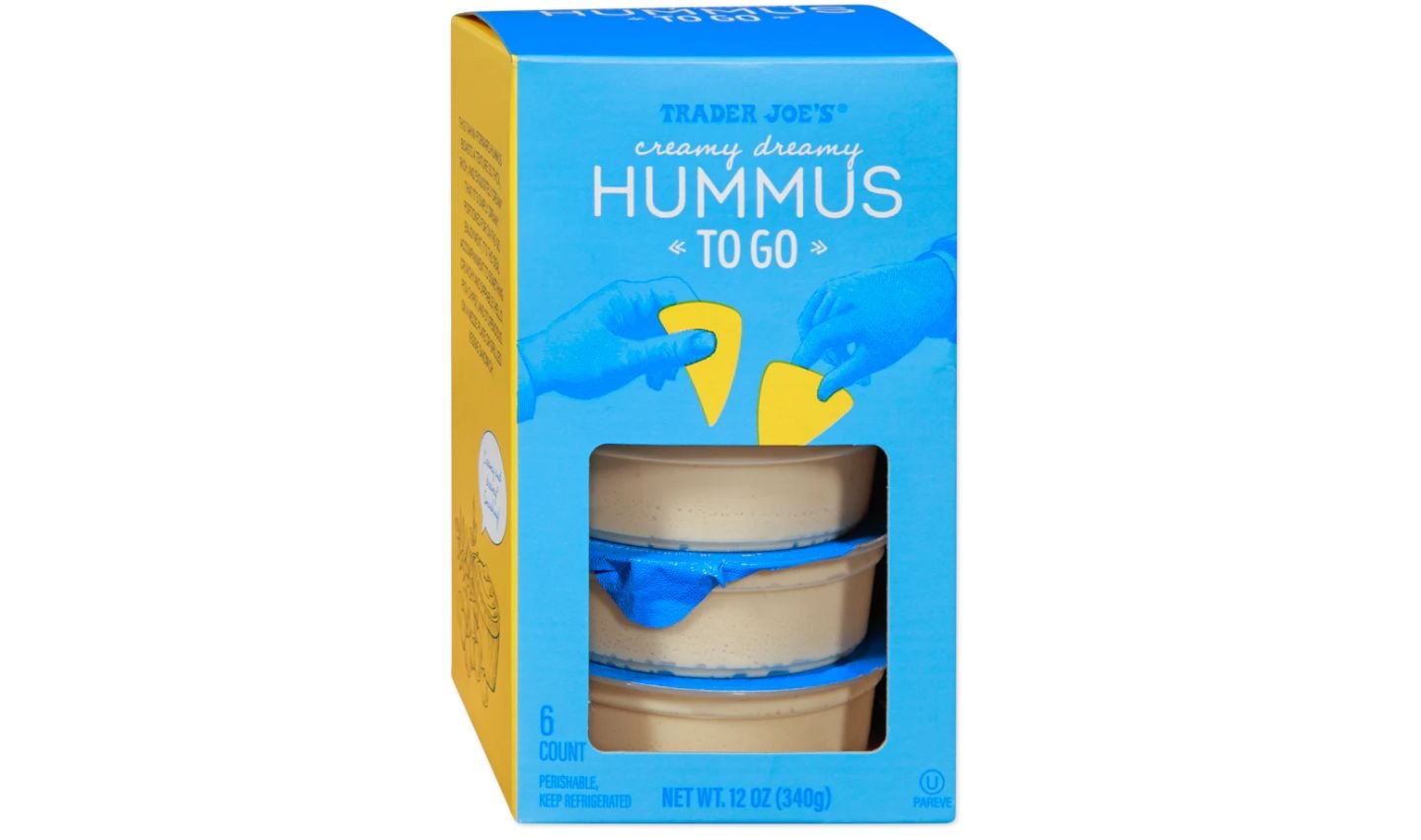 A box of Trader Joe’s Creamy Dreamy Hummus To Go, containing six individual hummus cups, is shown with blue and yellow packaging. The front of the box has a cutout revealing the hummus cups inside.