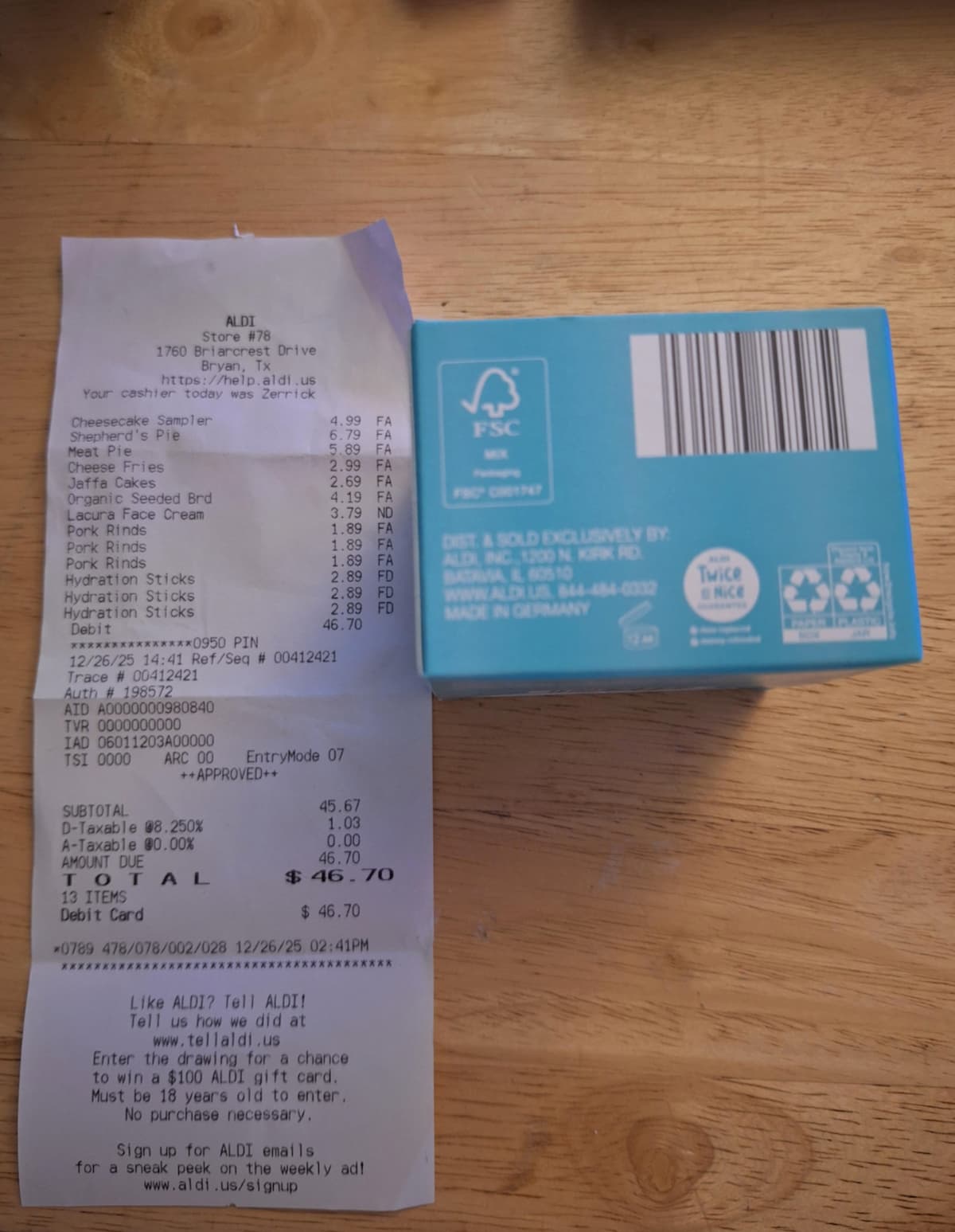 A receipt from ALDI with a total of $46.70 lies on a wooden table next to a blue food package showing recycling and FSC logos, a barcode, and product information.
