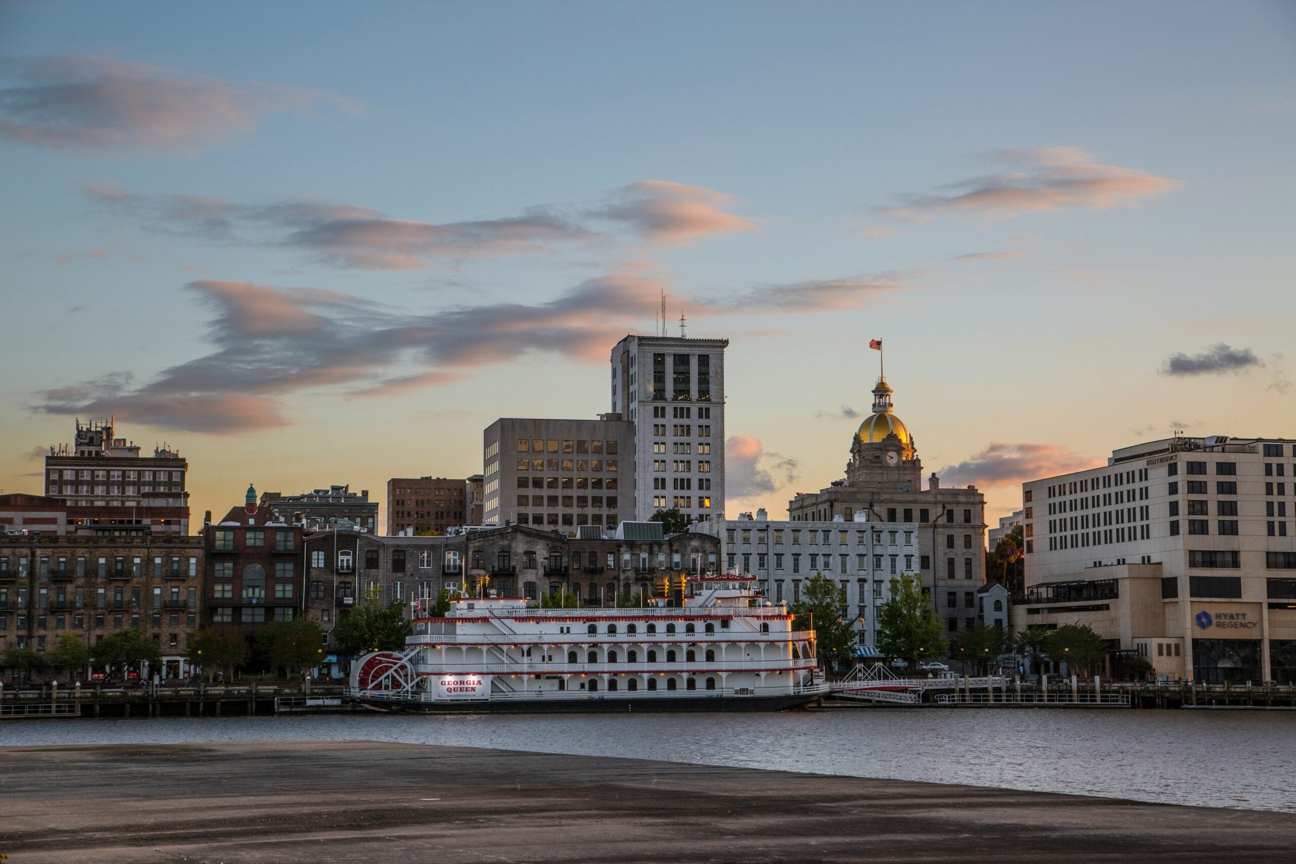 A riverboat with bright lights is docked along a waterfront, with historic and modern buildings, including a gold-domed structure, in the background at sunset under a partly cloudy sky.