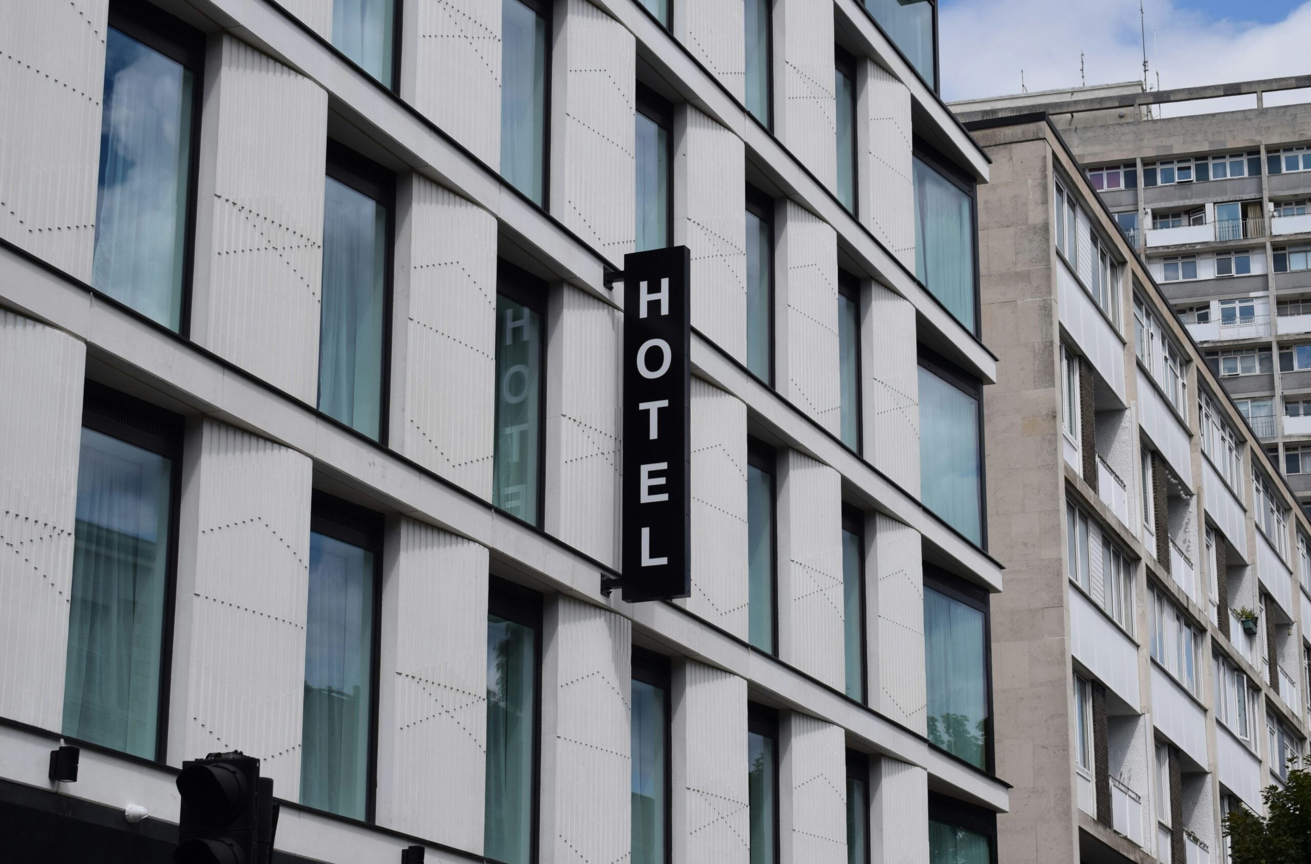 A modern white building with large windows has a vertical black sign reading "HOTEL" in bold white letters. Another tall building is visible in the background.