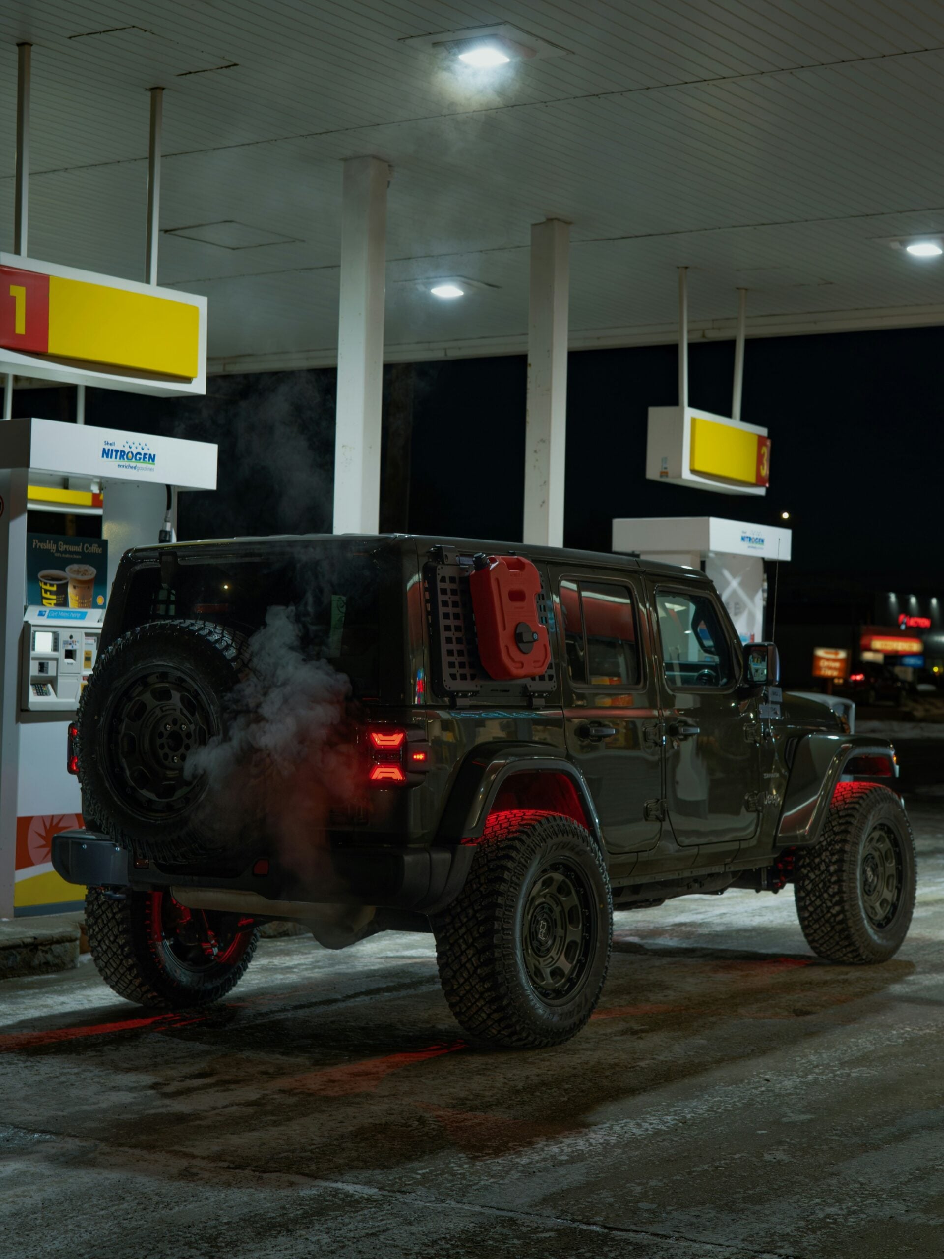 A black Jeep Wrangler with large tires is parked at a gas station at night, with exhaust steam visible from the tailpipe. The area is dimly lit by overhead lights from the fuel pumps.