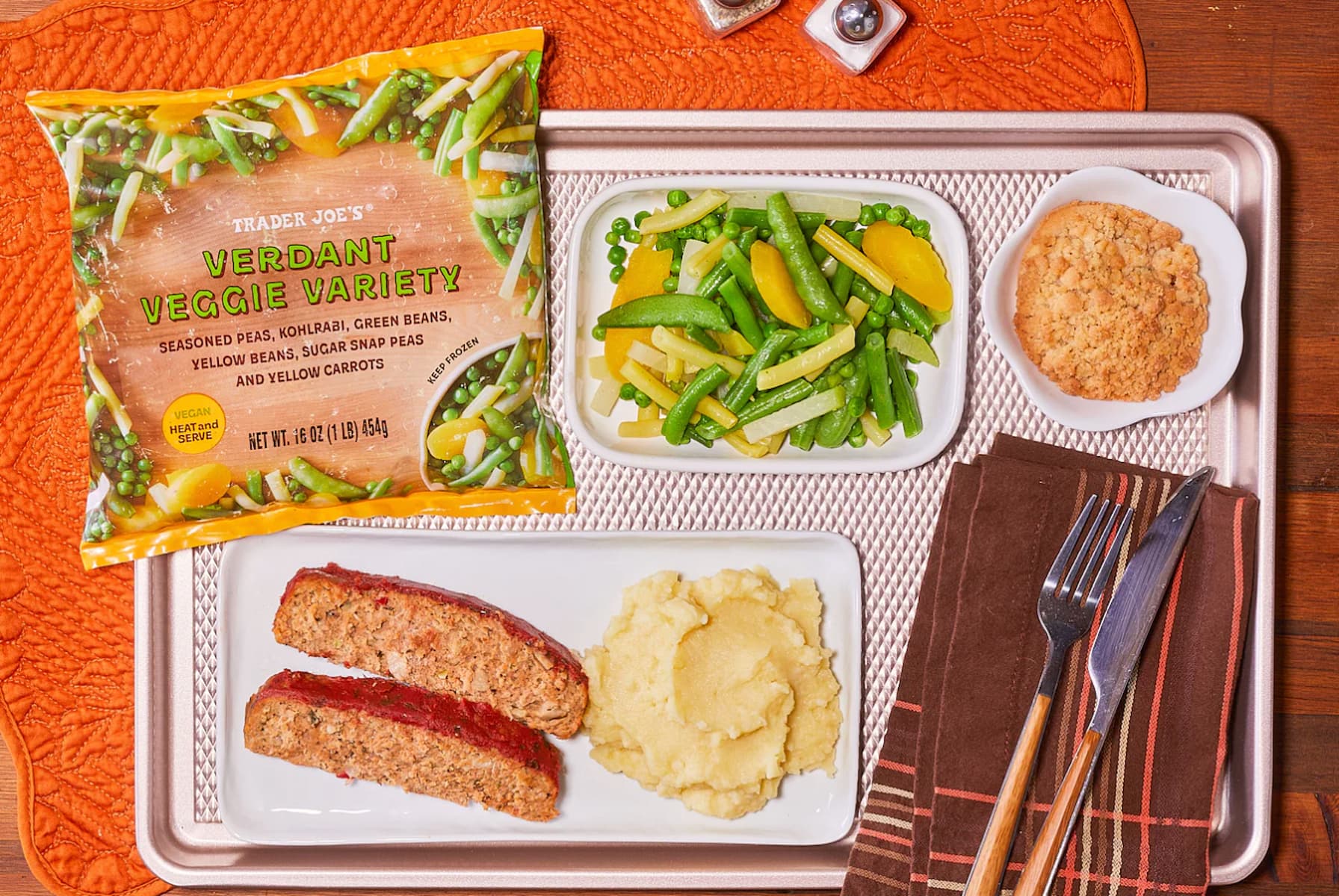 A tray with meatloaf, mashed potatoes, mixed veggies, a cookie, and a package of Trader Joe's Verdant Veggie Variety on a wooden table with an orange placemat, napkin, fork, and knife.