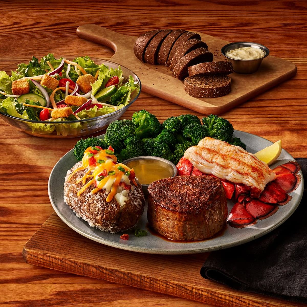 A plate with filet mignon, lobster tail, broccoli, loaded baked potato, and sauce. Nearby are a garden salad with croutons, rye bread slices, and a ramekin of butter, all on a wooden table.