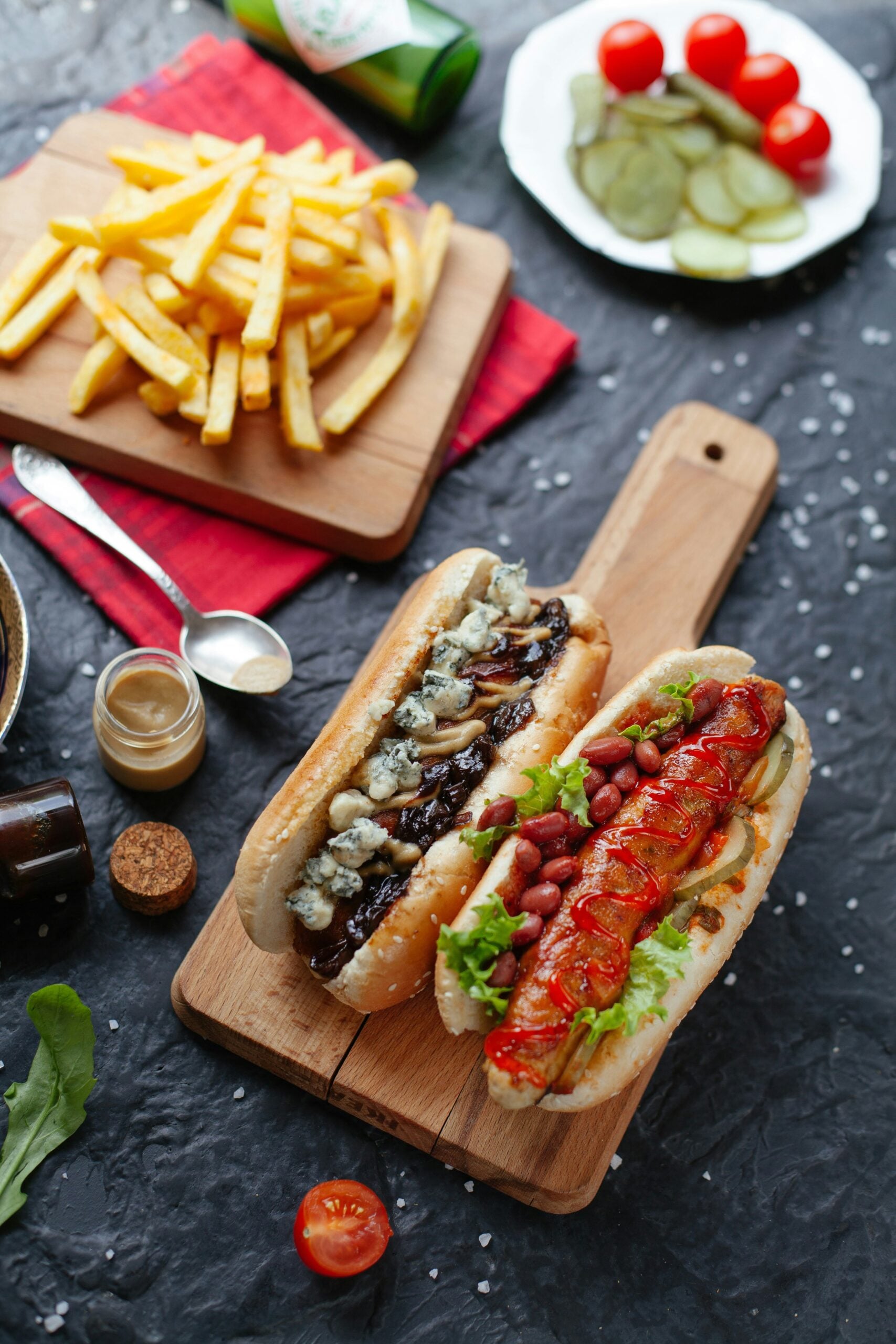 Two hot dogs on a wooden board; one topped with mushrooms and blue cheese, the other with lettuce, sausage slices, and sauce. French fries, pickles, cherry tomatoes, and condiments are nearby on a dark textured surface.