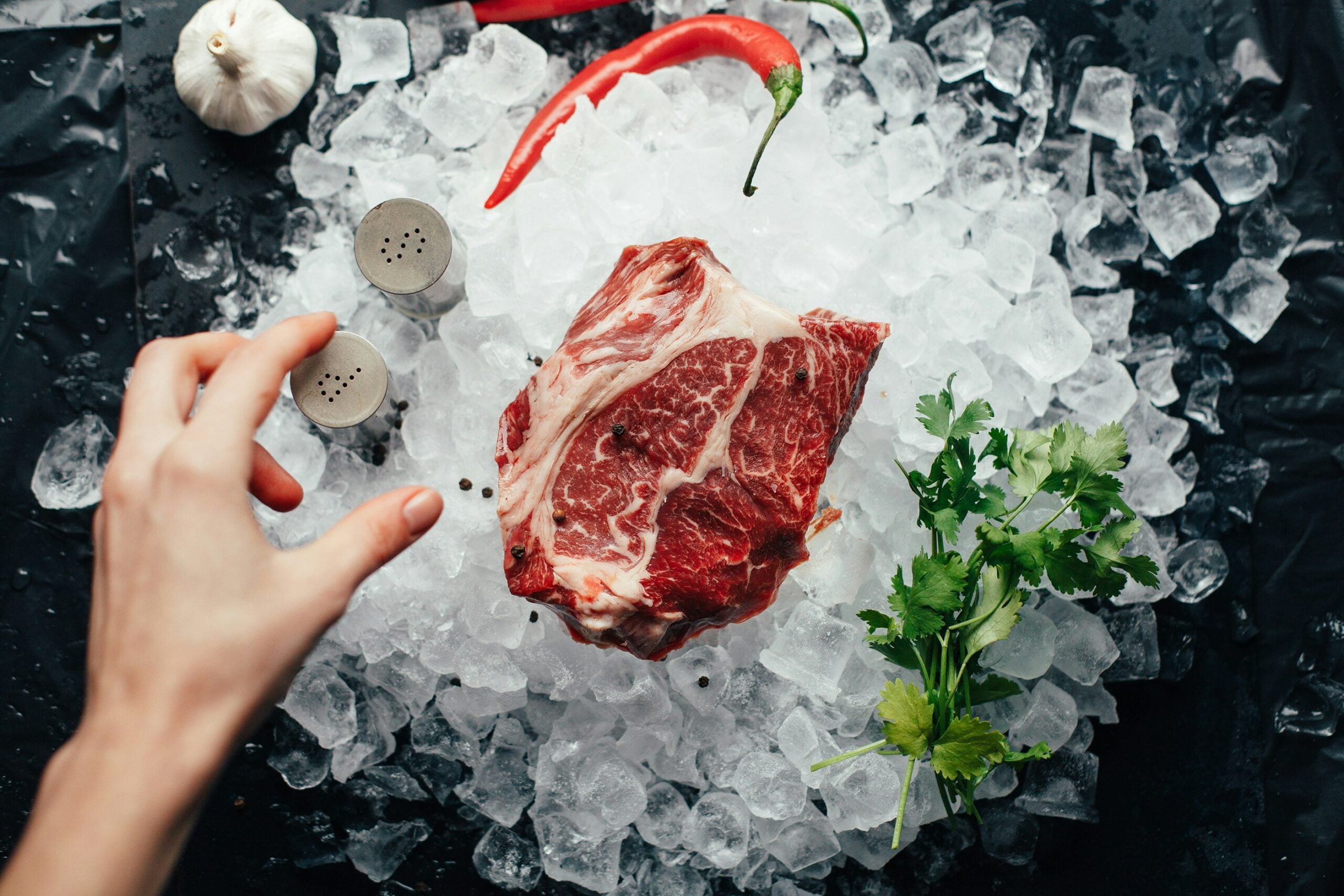 A raw marbled steak sits on a bed of ice, surrounded by fresh cilantro, a chili pepper, garlic, and salt and pepper shakers. A hand reaches toward the steak from the left side of the image.