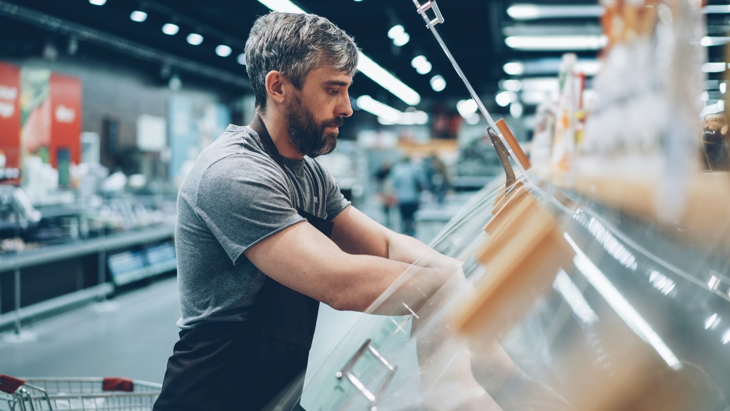 A man with short hair and a beard, wearing a gray shirt and black apron, works behind the counter at a grocery store deli, arranging or packaging items. Shopping carts and store shelves are visible in the background.
