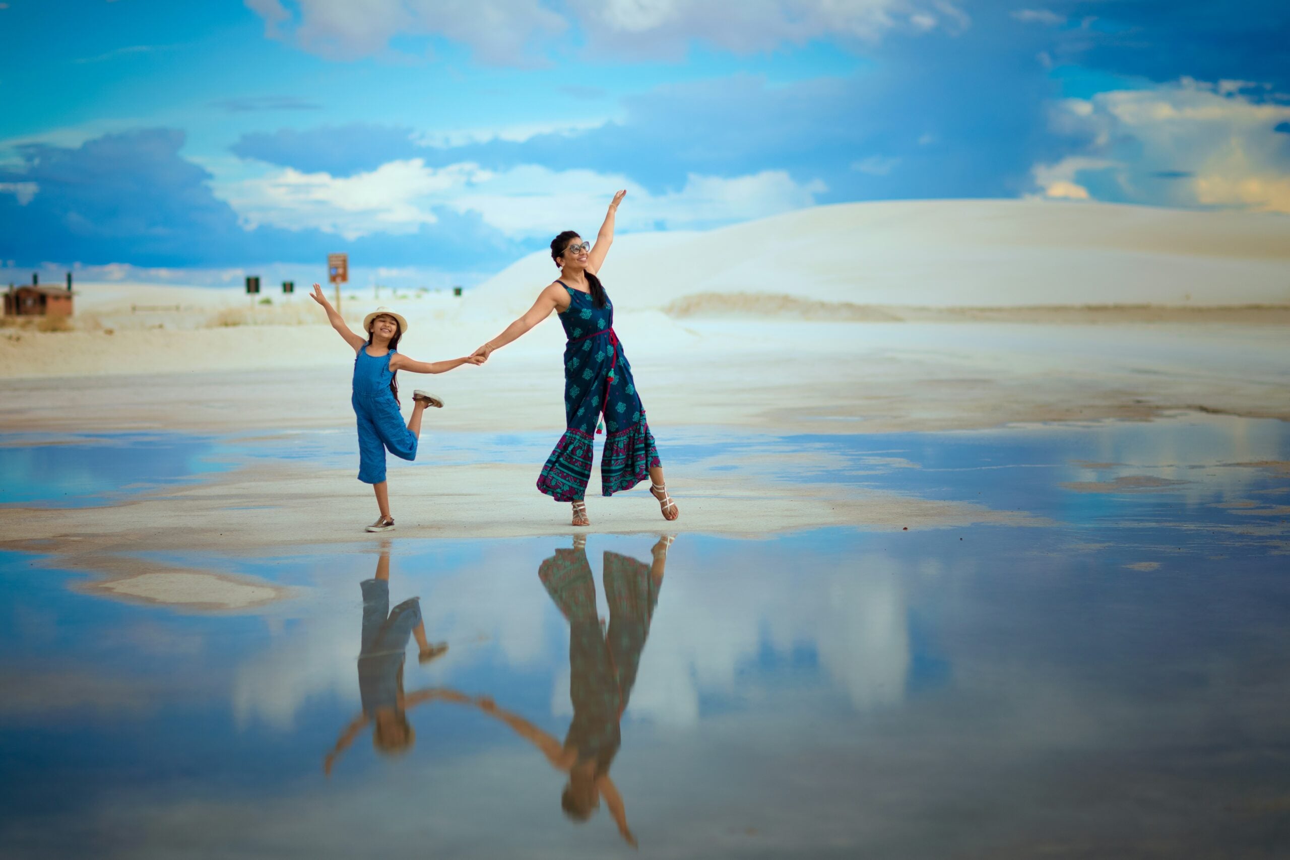 A woman and a child hold hands, dancing joyfully on a wet sandy surface with their reflections visible. The sky is blue with clouds, and white sand dunes are in the background.