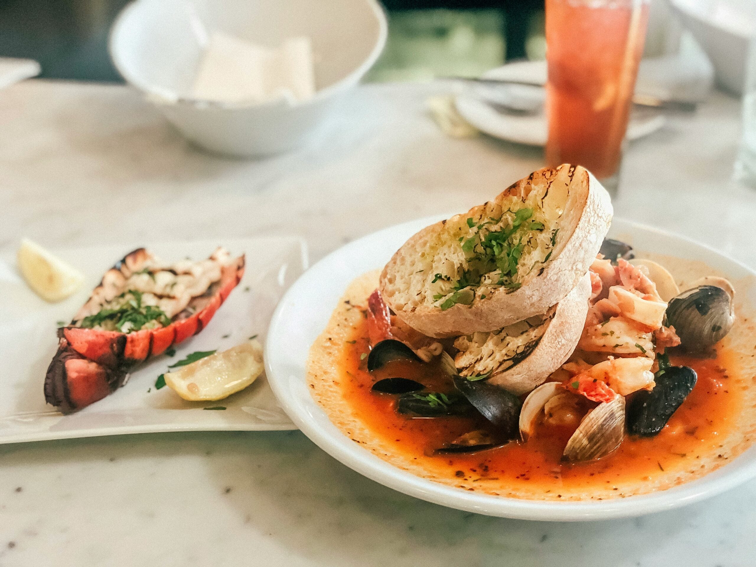 A seafood stew with clams, mussels, and bread slices is served in a white bowl, with a lobster tail and a lemon wedge on a separate plate in the background.