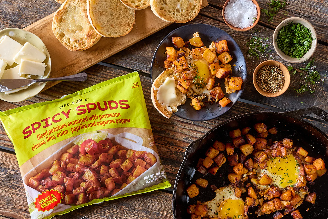A bag of Trader Joe’s Spicy Spuds is on a table with cooked potatoes in a skillet and on a plate with eggs. Surrounding items include sliced bread, butter, chopped herbs, salt, and pepper.