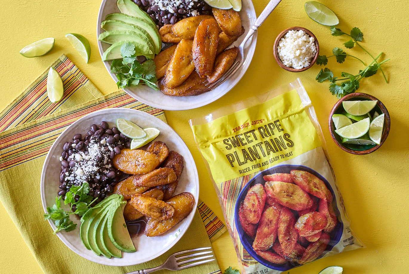 Two plates with sliced avocado, sweet plantains, black beans topped with cheese, cilantro, and lime wedges. A striped napkin, bowls of cheese, lime, and a bag of Trader Joe’s Sweet Ripe Plantains are nearby on a yellow surface.