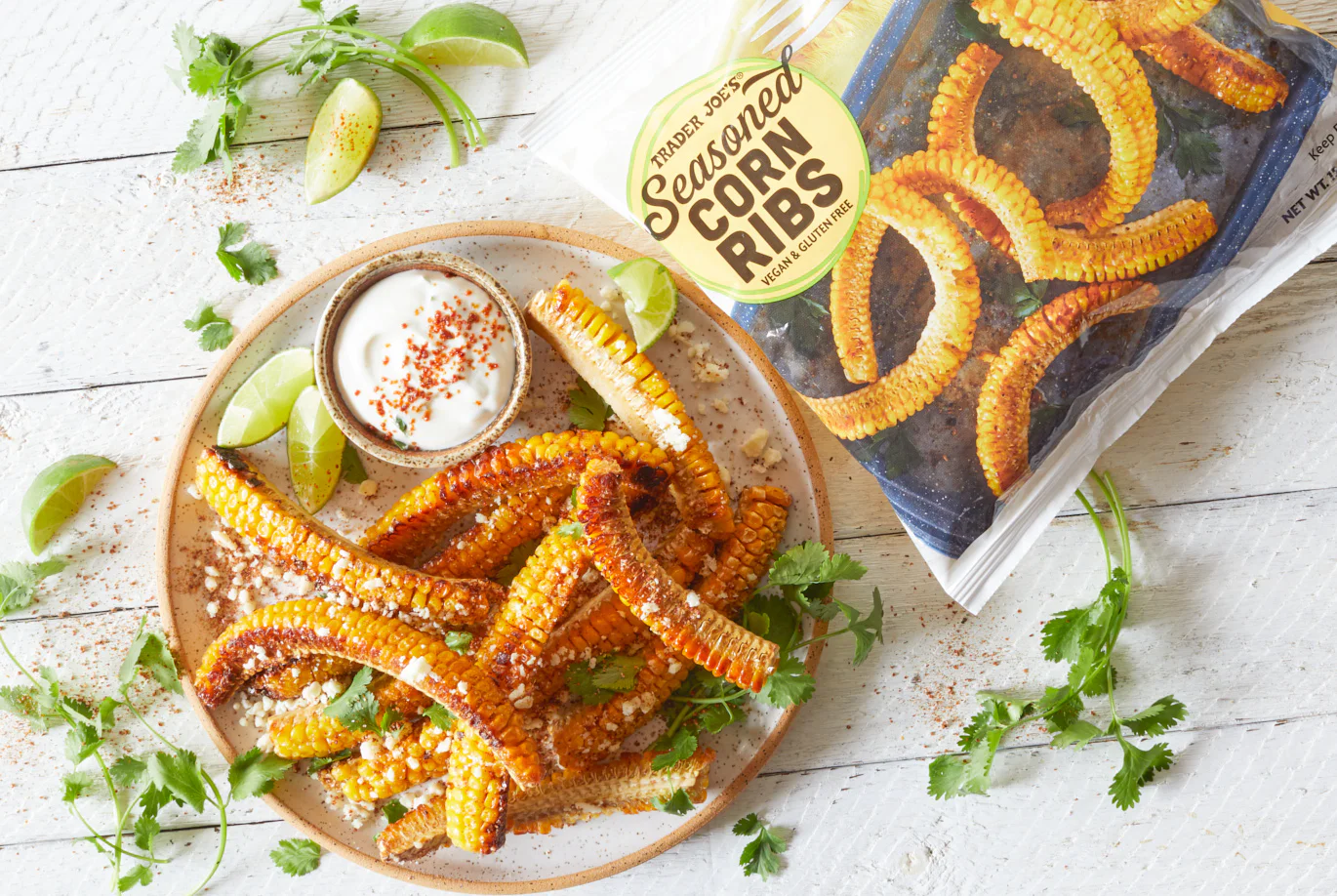 A plate of roasted corn ribs sprinkled with cheese and spices, served with a bowl of dipping sauce and lime wedges. Fresh cilantro and a package labeled “Seasoned Corn Ribs” are on a white wooden surface.