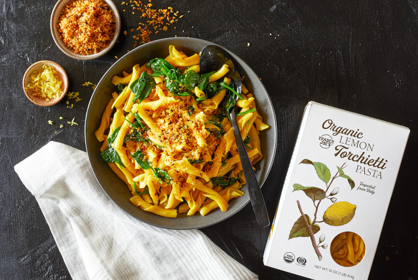 A bowl of pasta with spinach and breadcrumbs sits next to a box labeled "Organic Lemon Torchetti Pasta" on a dark surface, with a spoon, a small bowl of breadcrumbs, and a striped napkin nearby.