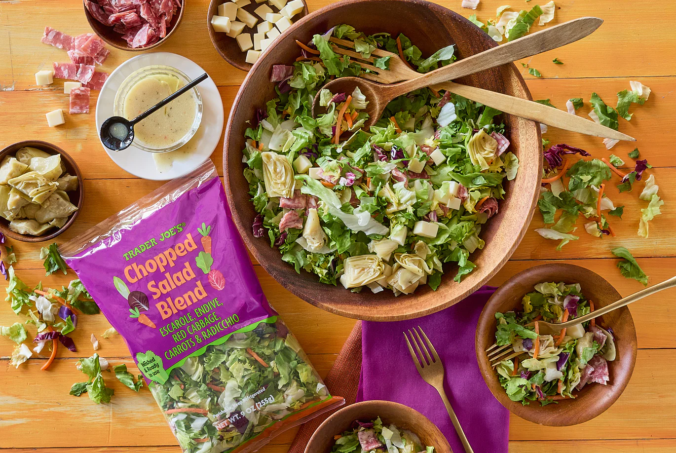 A wooden bowl of chopped salad with artichoke hearts, cheese cubes, and salami on a wooden table, surrounded by smaller bowls of salad, a packet of salad blend, dressing, and utensils.
