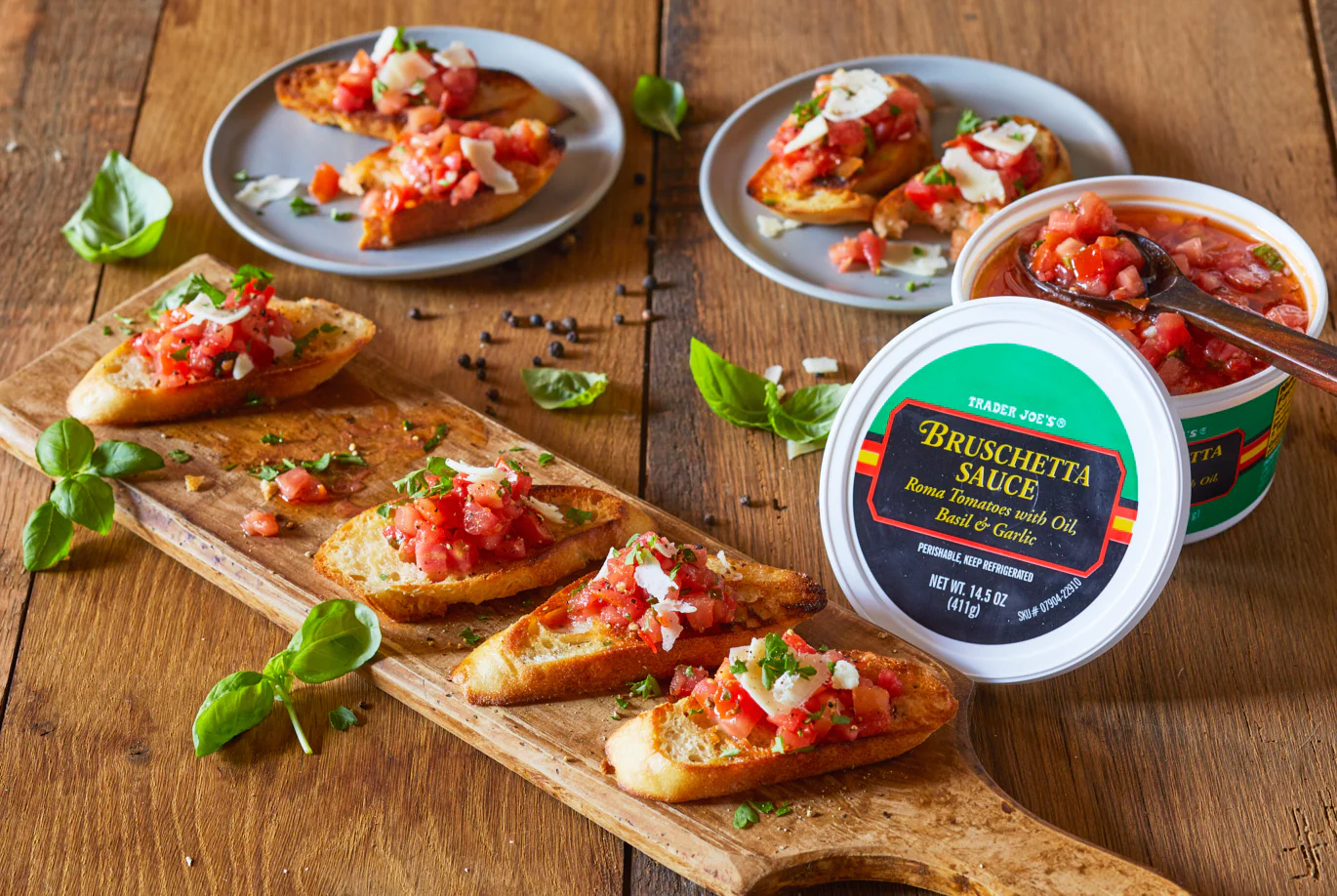 A wooden board with toasted bread topped with bruschetta sauce and cheese, garnished with herbs. Two small plates with bruschetta and an open container of Trader Joe's Bruschetta Sauce are also on the wooden table.