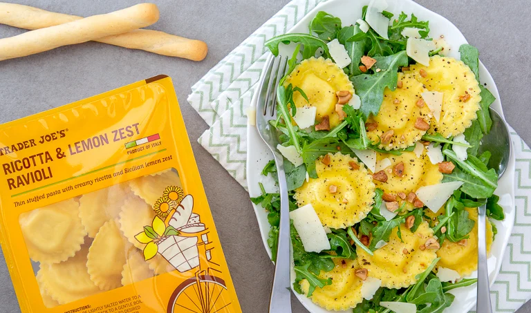A plate of ravioli with arugula, shaved cheese, and nuts is next to a yellow package labeled “Trader Joe’s Ricotta & Lemon Zest Ravioli.” Two breadsticks and a patterned napkin are also visible.