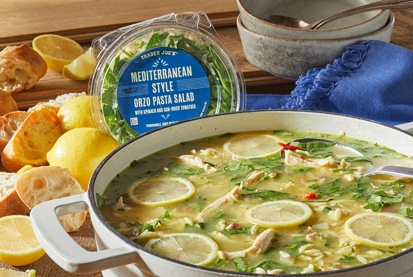 A pot of soup with lemon slices, chicken, and herbs sits beside a container of Trader Joe’s Mediterranean Style Orzo Pasta Salad, sliced bread, lemons, and a blue napkin on a wooden table.