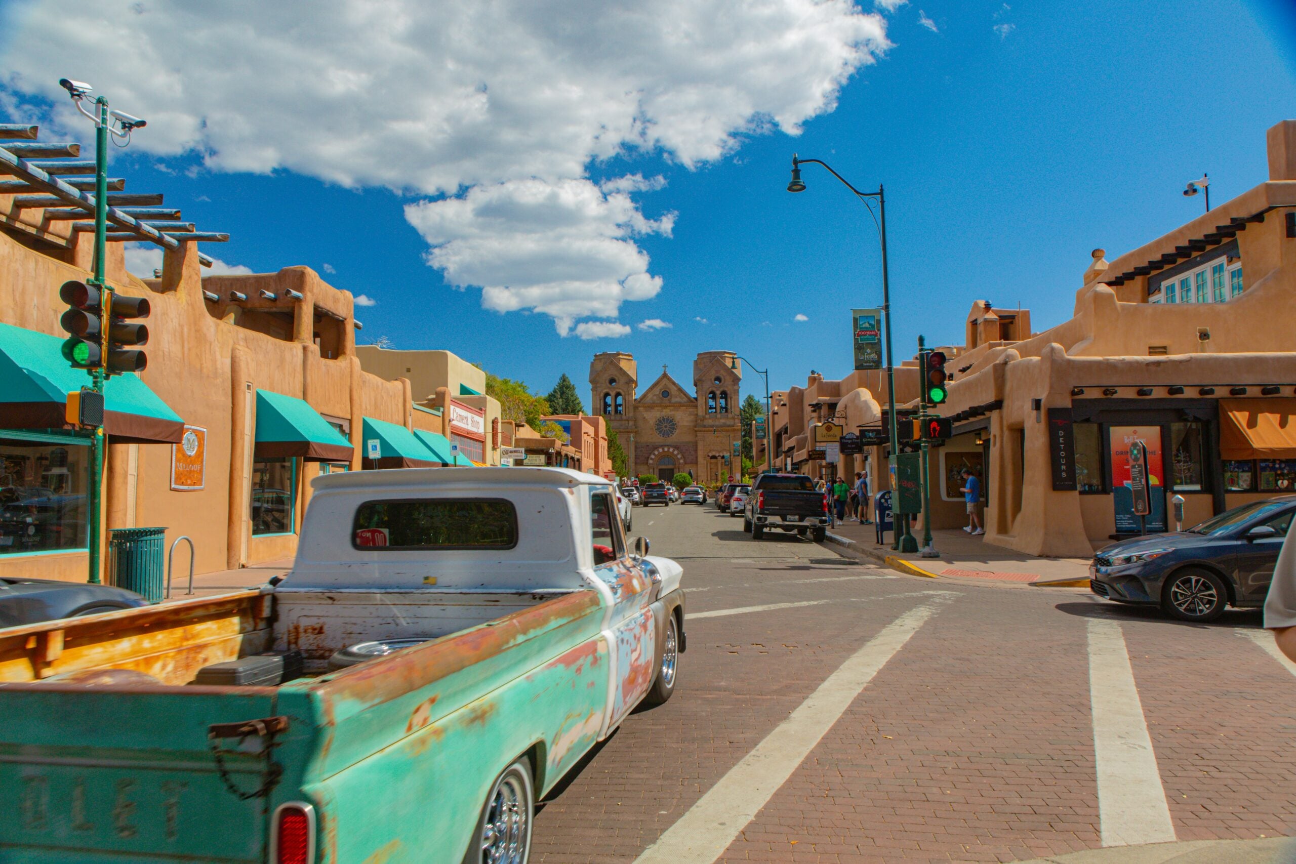 A weathered vintage pickup truck drives down a busy street lined with adobe-style buildings and shops under a bright blue sky with fluffy clouds, in a southwestern town.
