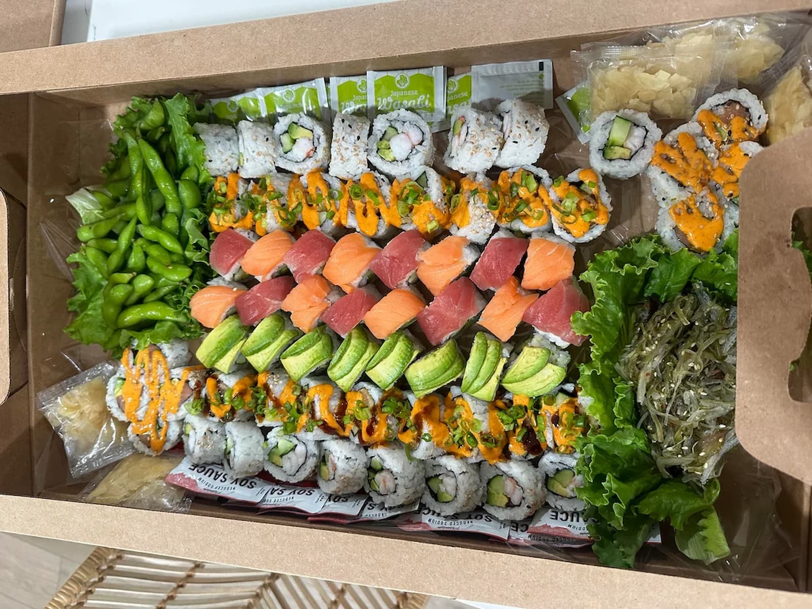 A large sushi platter in a cardboard box filled with various rolls, edamame, avocado, tuna, salmon, and seaweed salad.