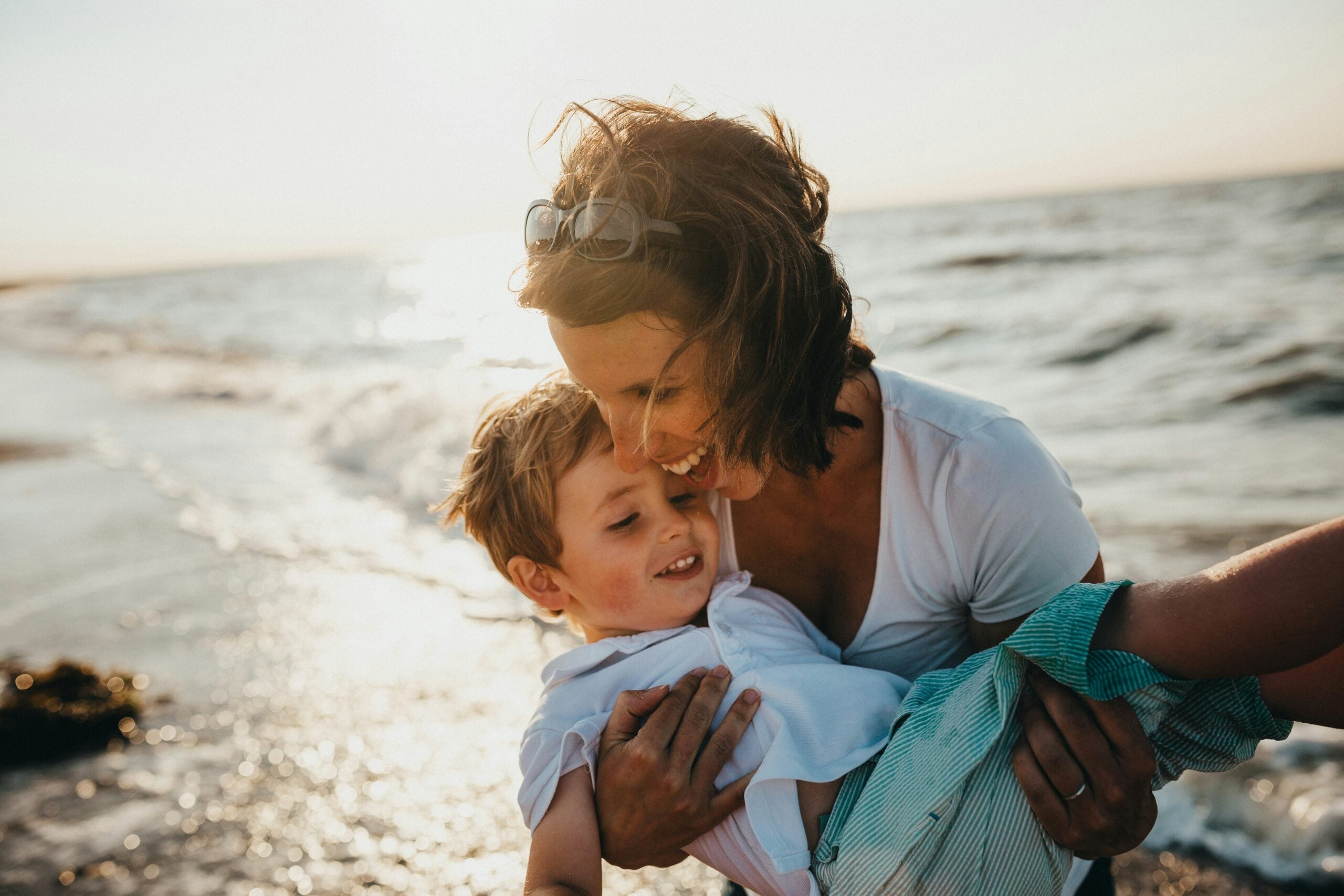 A woman smiles and hugs a young boy while standing on a beach at sunset. The ocean waves are in the background, and both appear joyful and happy.
