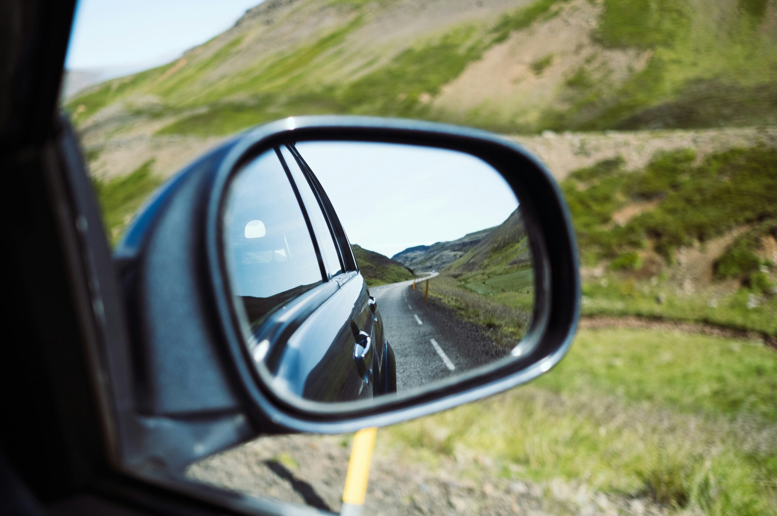 A car’s side mirror reflects a winding road and green hills, showing the landscape behind the vehicle. The surrounding scenery is lush and mountainous, with clear skies and vibrant vegetation.