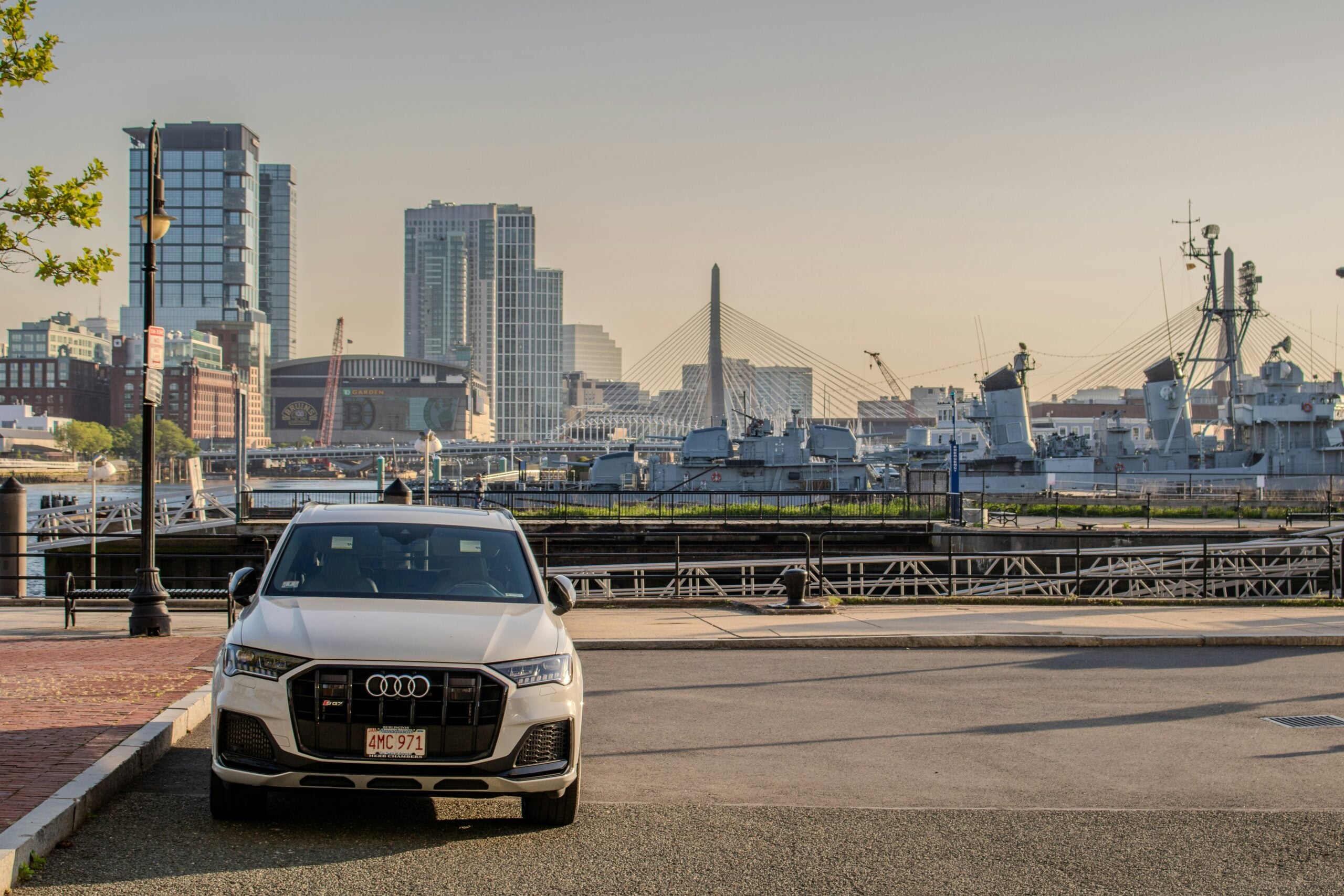 A white Audi SUV is parked near a harbor with battleships, city buildings, and a cable-stayed bridge visible in the background under a clear sky.