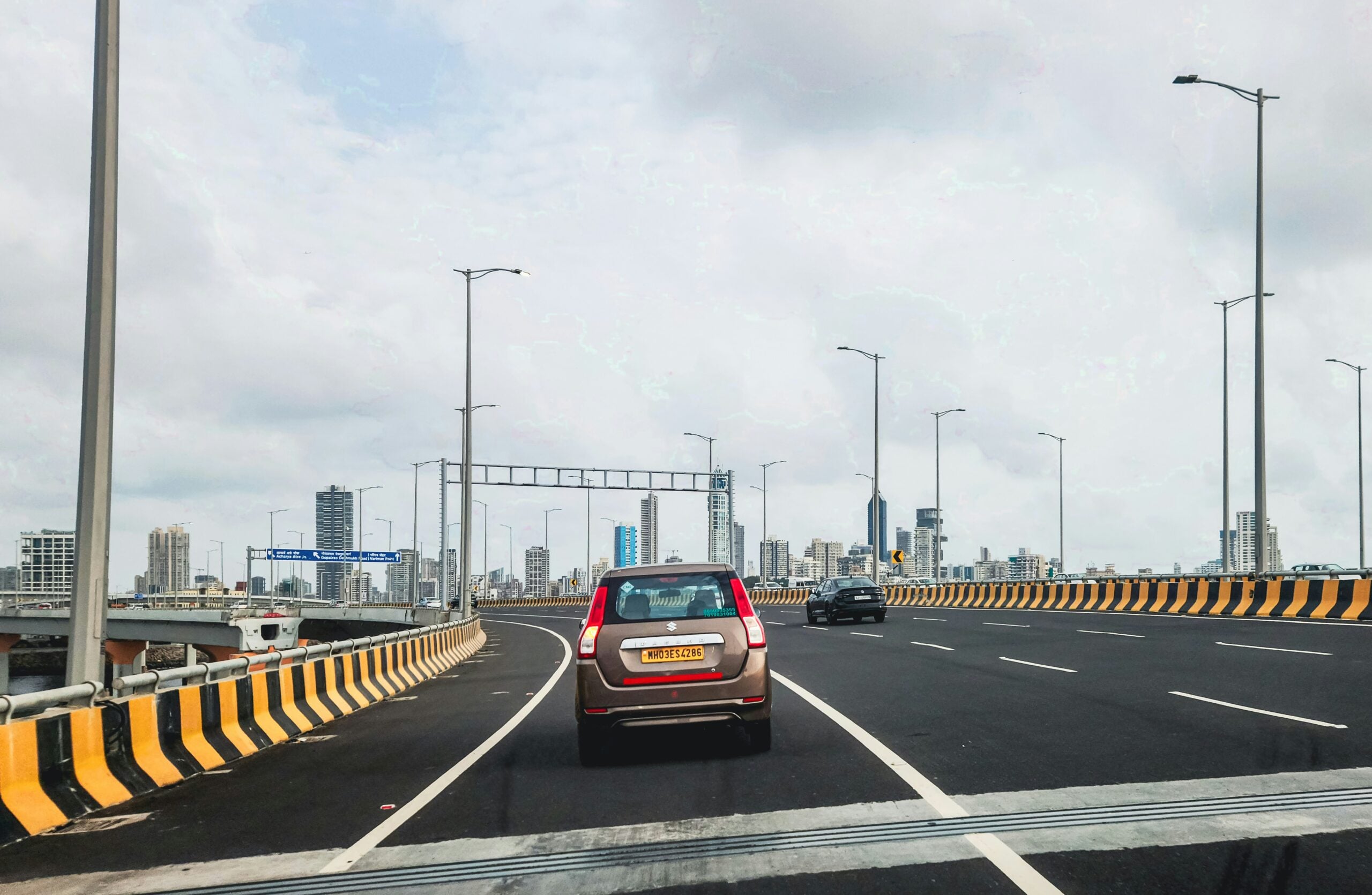 A brown car drives on a modern, elevated road with yellow and black barriers. Tall streetlights line the road, and a city skyline with high-rise buildings is visible under a cloudy sky.
