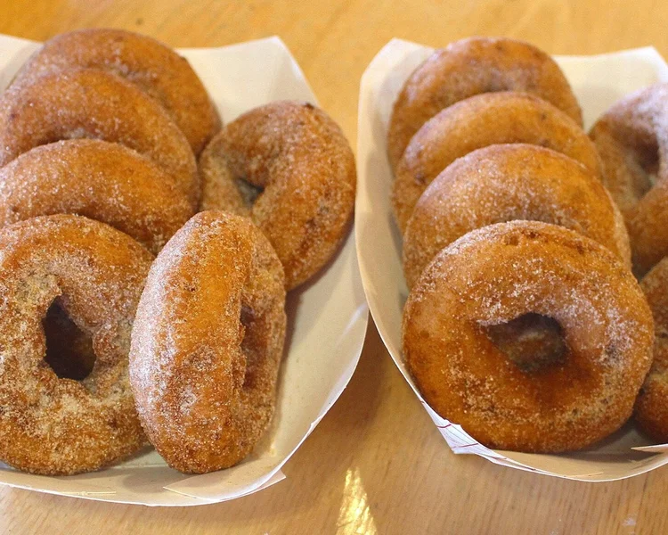 Two paper trays filled with fresh, sugar-coated apple cider donuts sit on a wooden surface, with the donuts golden brown in color and dusted with a layer of granulated sugar.