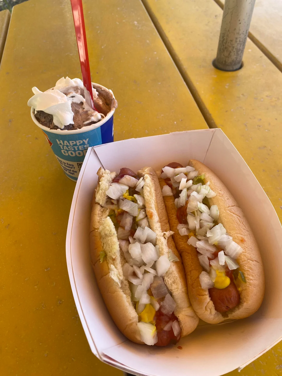 Two Fast-Food Hot Dogs topped with chopped onions, relish, ketchup, and mustard in a paper tray sit beside a cup of ice cream with a red spoon, all on a yellow outdoor picnic table.