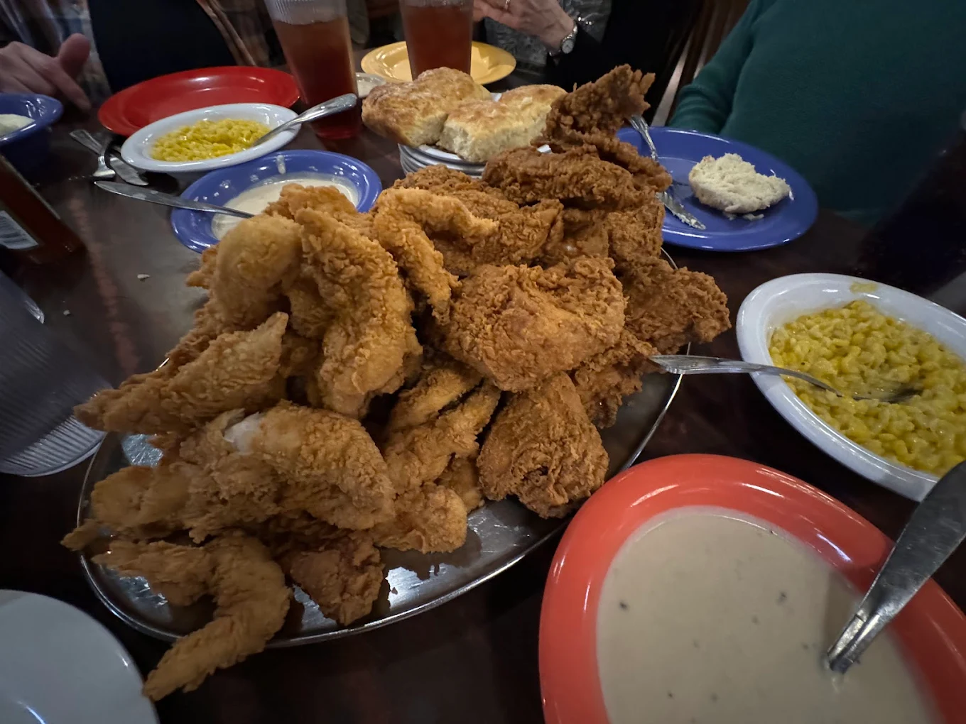 A large platter of fried chicken is surrounded by plates of corn, biscuits, mashed potatoes, and drinks on a table, with several people seated around it.