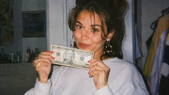 A woman with long brown hair in a white top holds a ten-dollar bill and smiles at the camera, perhaps reflecting on 1991 prices. She is indoors, with shelves and clothes visible in the background.