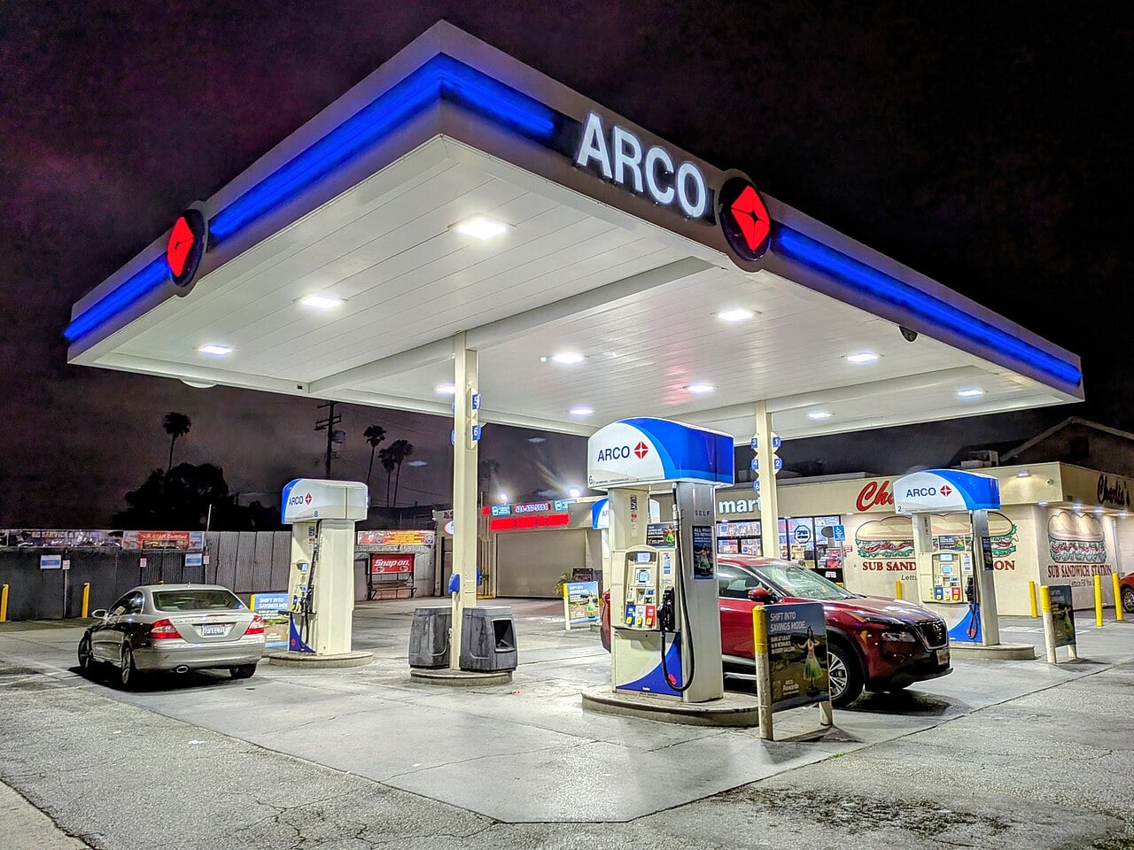 A brightly lit ARCO gas station at night with two cars parked at separate fuel pumps; the convenience store is visible in the background under a dark sky.