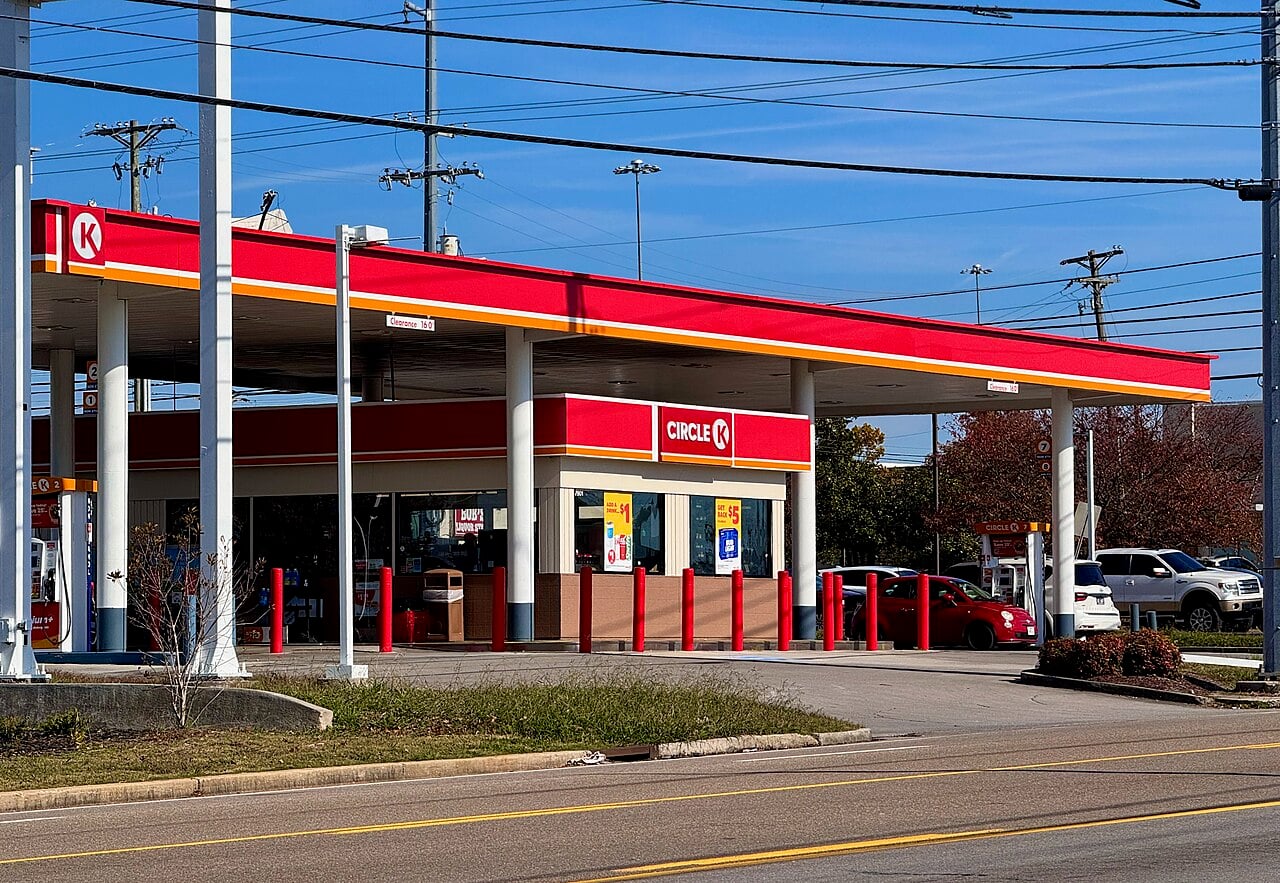 A Circle K gas station with red and yellow signage, multiple fuel pumps, and several cars parked nearby under a clear blue sky. Power lines and a few trees are visible in the background.